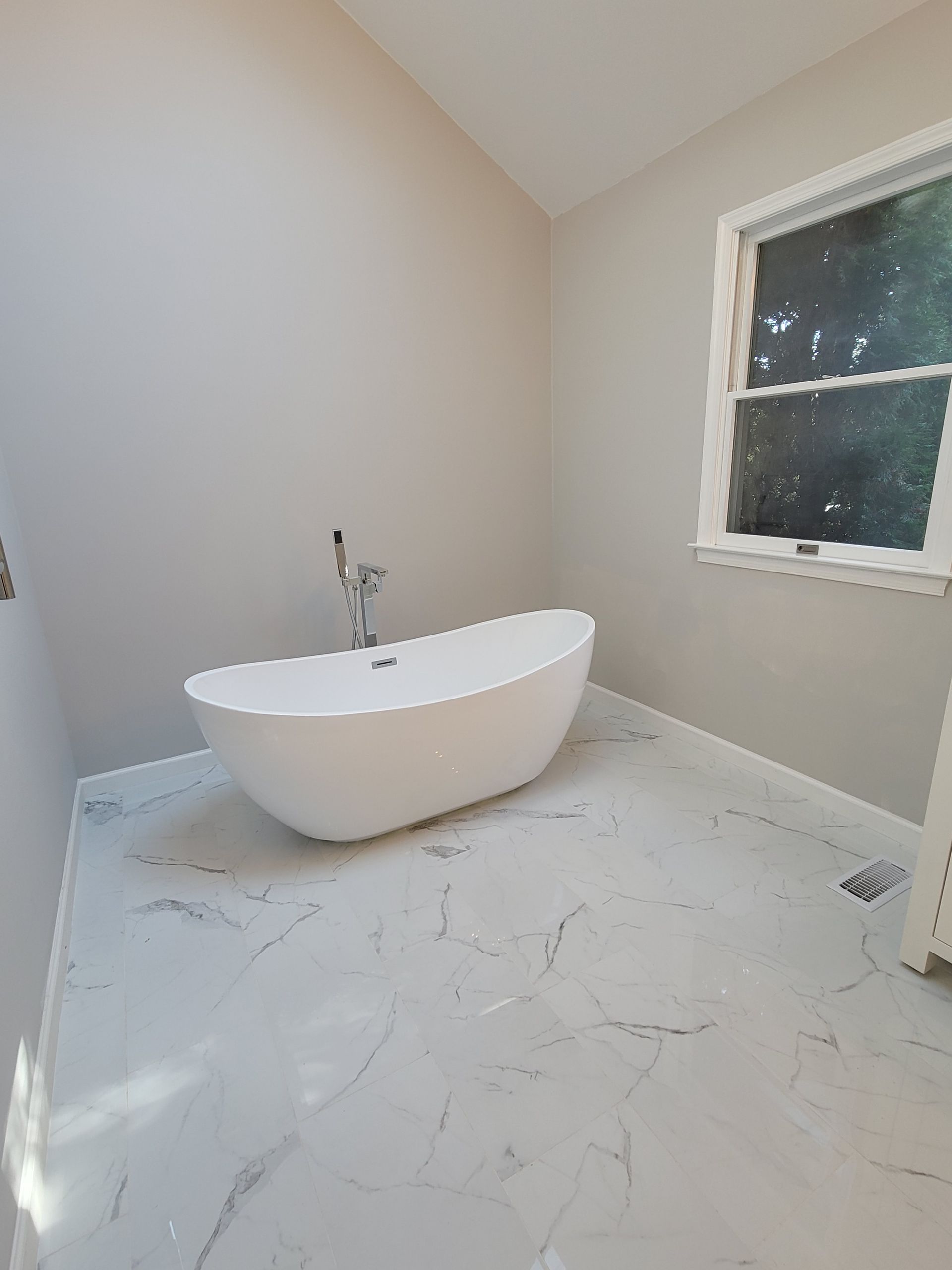 A white freestanding bathtub sits on a white marble-look floor in a room with light gray walls and a single window.