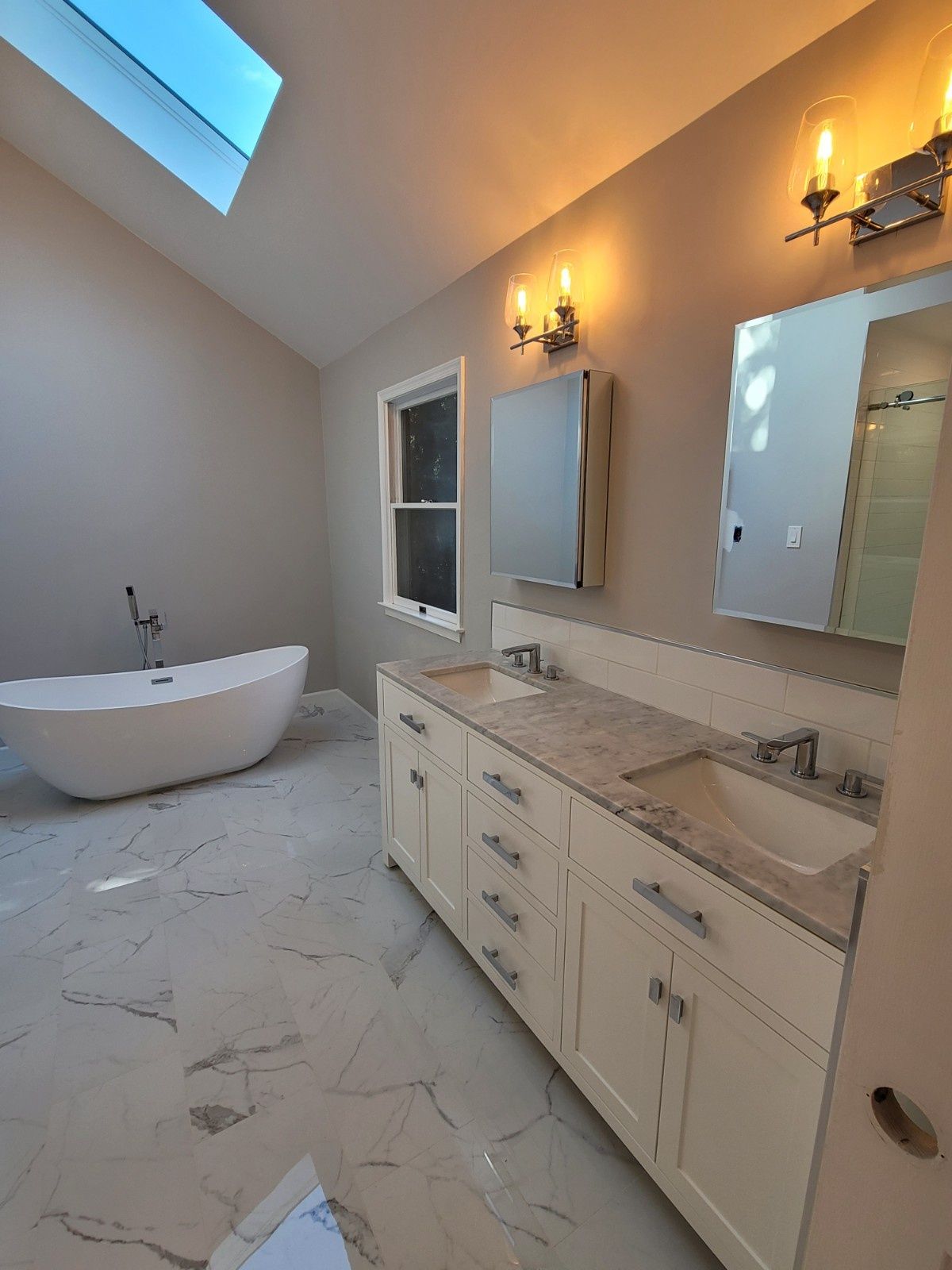 A modern bathroom featuring a white freestanding tub, double vanity with stone countertop, and marble-look floor tiles.