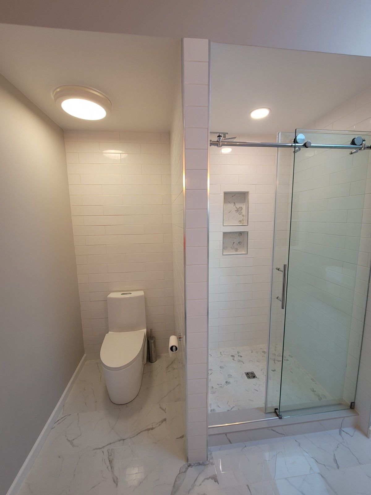 A modern bathroom with white marble-style flooring, a white toilet, and a glass-enclosed shower with tiled walls.