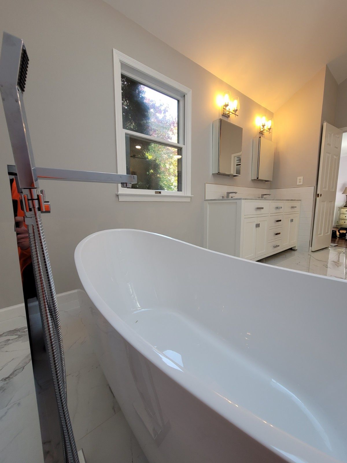 A freestanding white bathtub sits on a tiled floor in front of a window and a double vanity in a bright bathroom.
