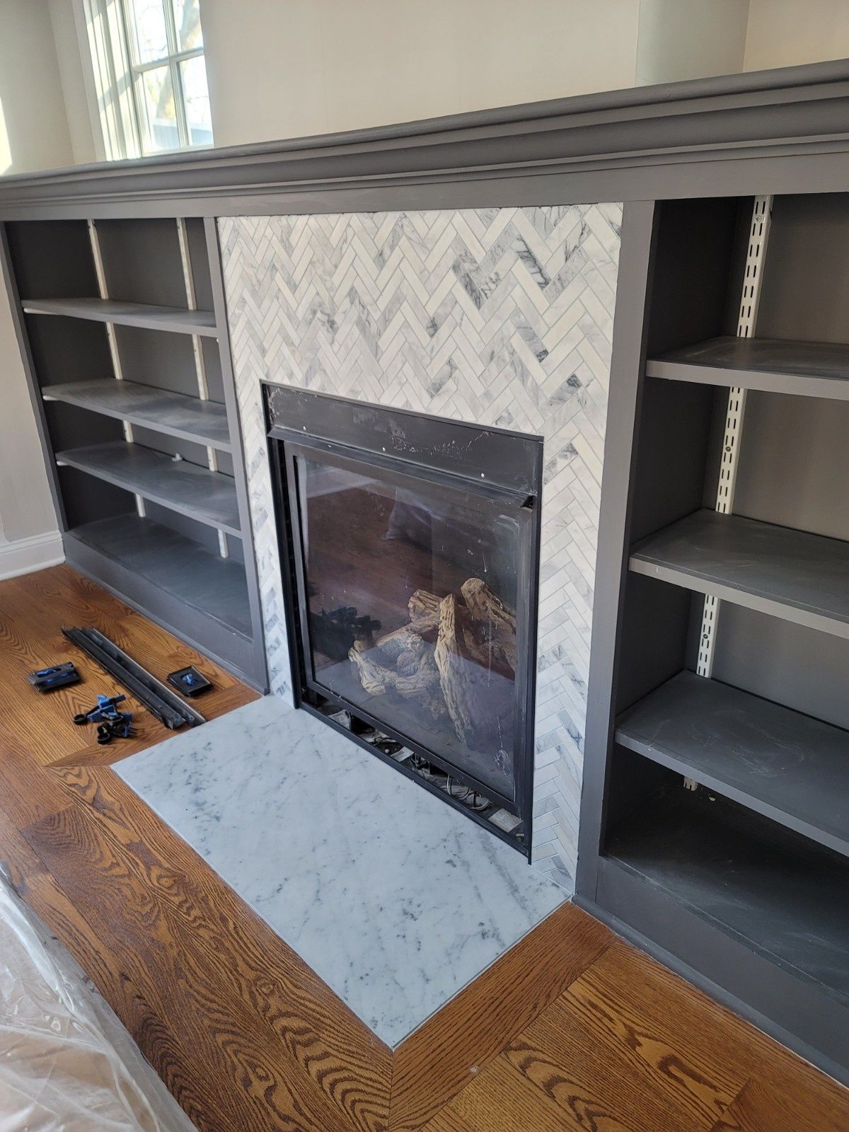 A fireplace with a white marble herringbone tile surround and hearth, flanked by dark gray built-in shelving.