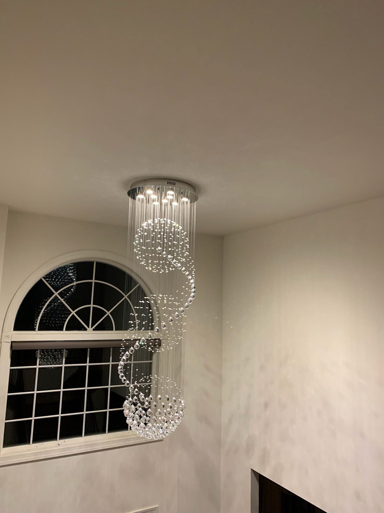 A spiral crystal chandelier hangs from a ceiling next to an arched window in a brightly lit room.