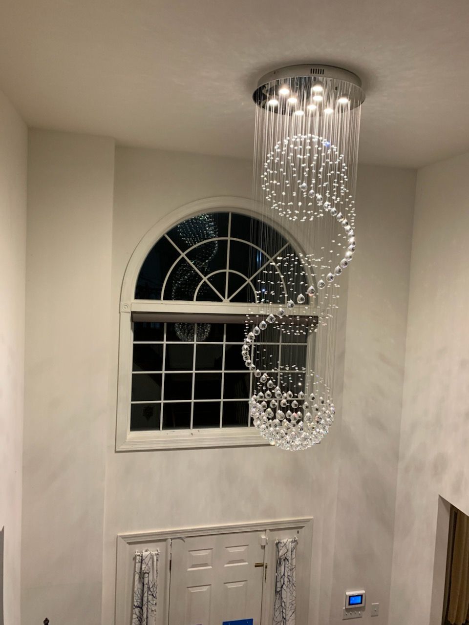 A modern, spiral crystal chandelier hangs from a high ceiling in a tall entryway with an arched window and white walls.