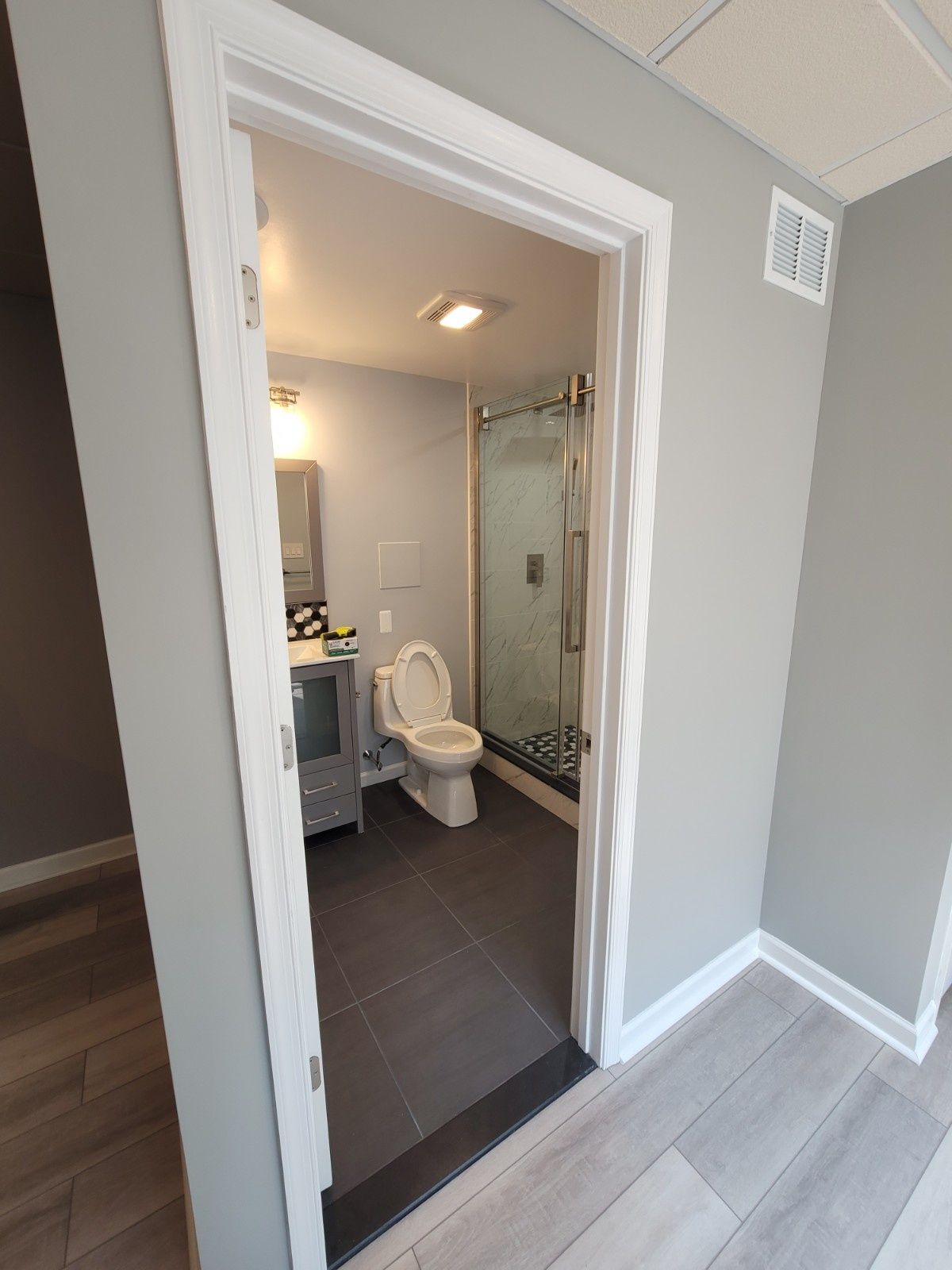 A doorway view of a small bathroom featuring a white toilet, grey vanity, glass-enclosed shower, and dark tile floor.