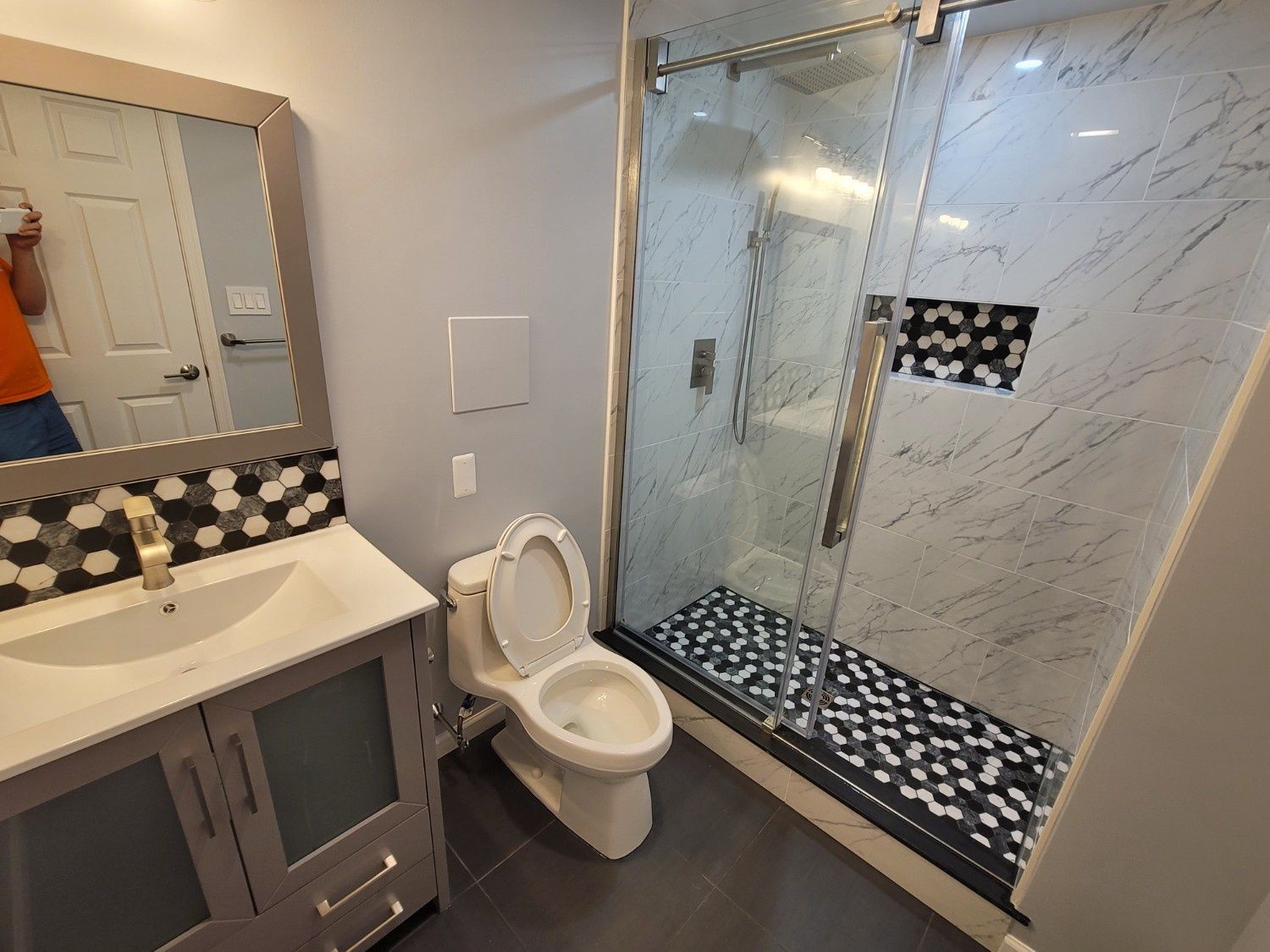 A modern bathroom with a grey vanity, a toilet, and a glass-enclosed shower featuring marble-look tiles and a mosaic floor.
