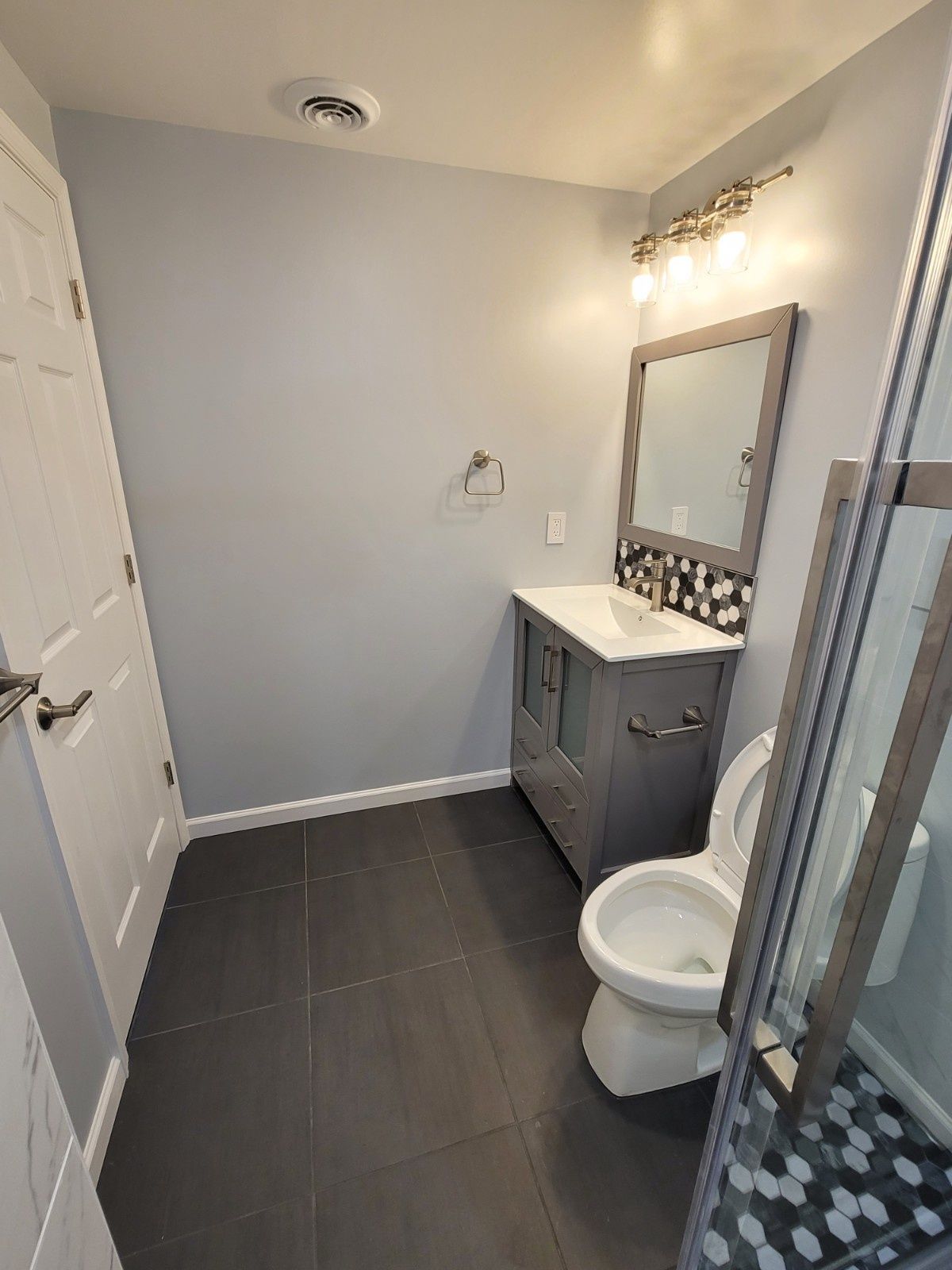 A small bathroom with light gray walls, dark gray floor tiles, a gray vanity with a mirror, and a white toilet.