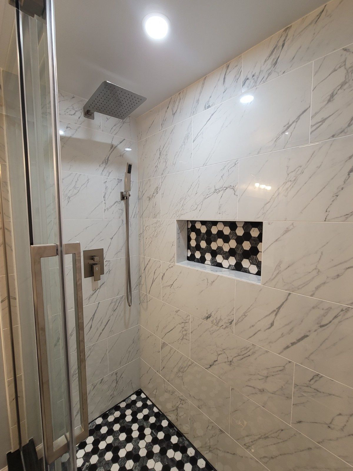 A modern shower with marble-look wall tiles, a recessed shelf, and a floor featuring black and white hexagonal tiles.