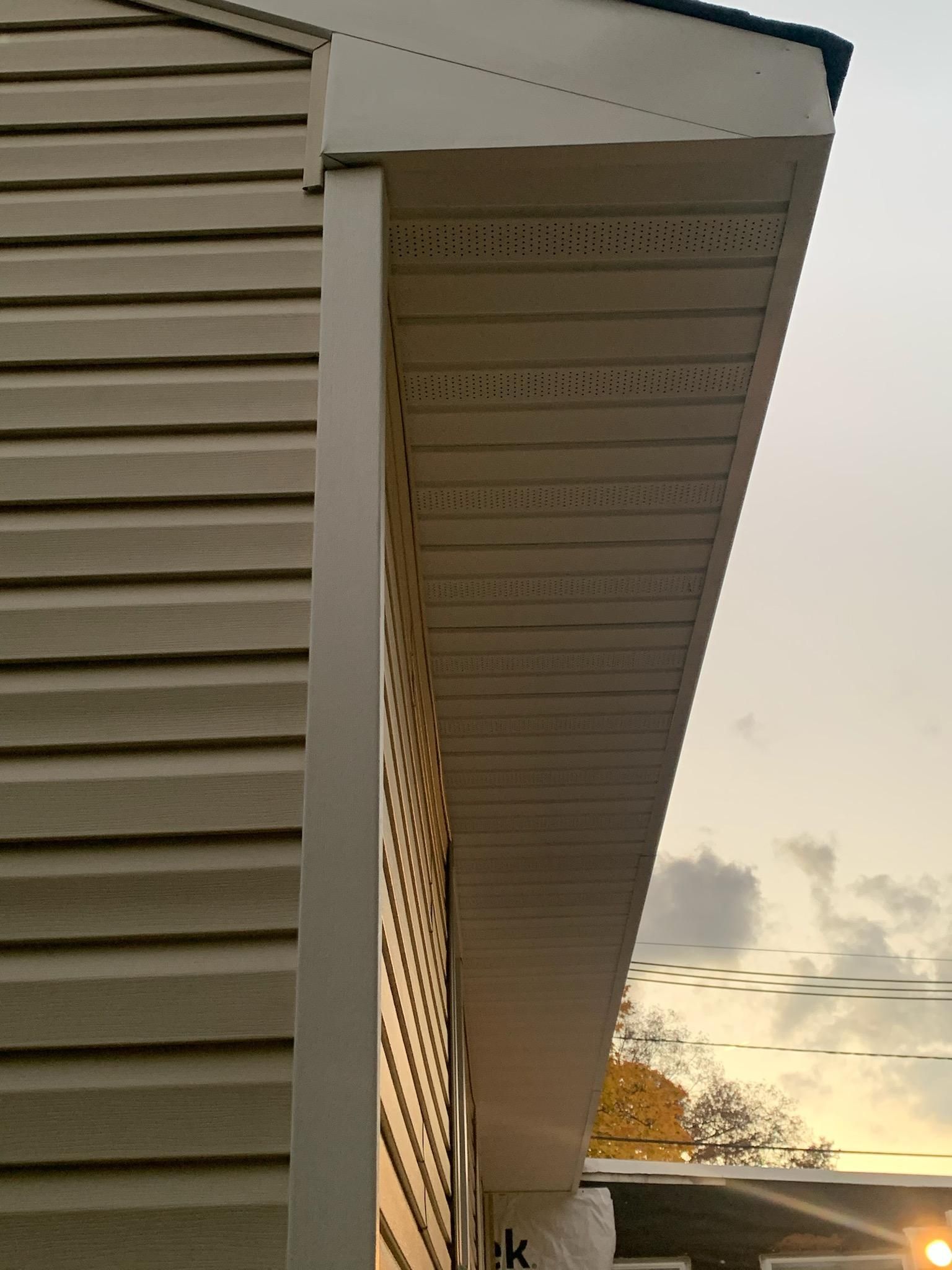 Close-up view of the exterior corner of a house showing beige vinyl siding, a white corner trim, and a vented soffit.