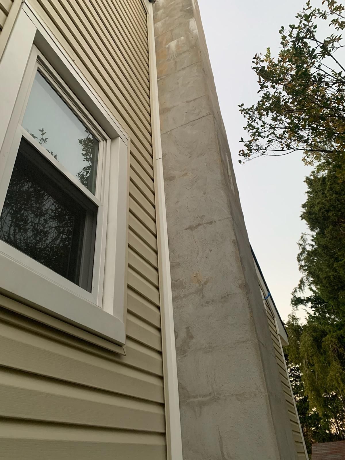 A close-up view of a light-colored house corner, featuring tan siding next to a vertical concrete chimney wall and window.