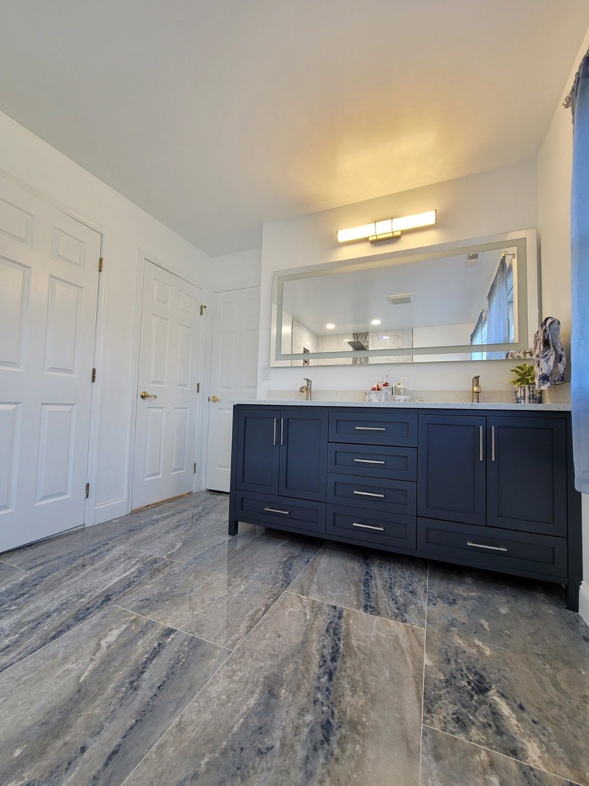 A modern bathroom featuring a dark blue double vanity with a wide mirror, white walls, and gray marble-patterned flooring.