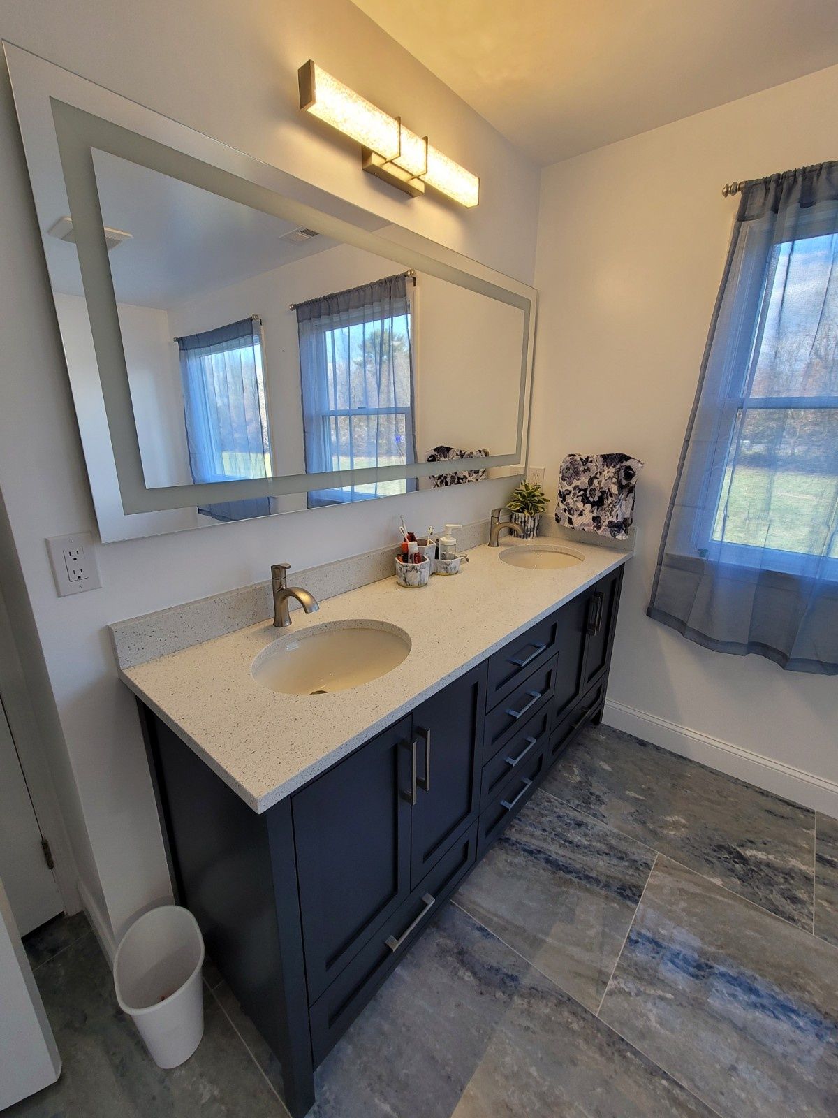 A bathroom vanity with a dark cabinet, speckled countertop, sink, and large mirror, with tiled floors and a window.