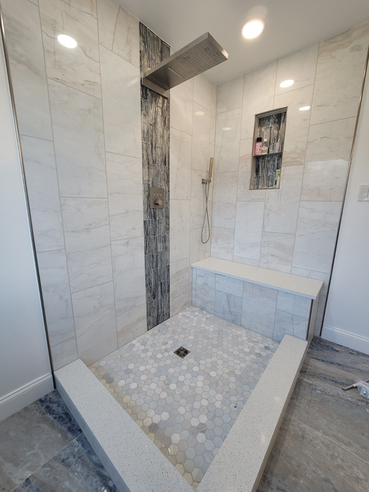 A walk-in shower with light marble-style wall tiles, a dark stone accent strip, a corner bench, and mosaic floor tile.
