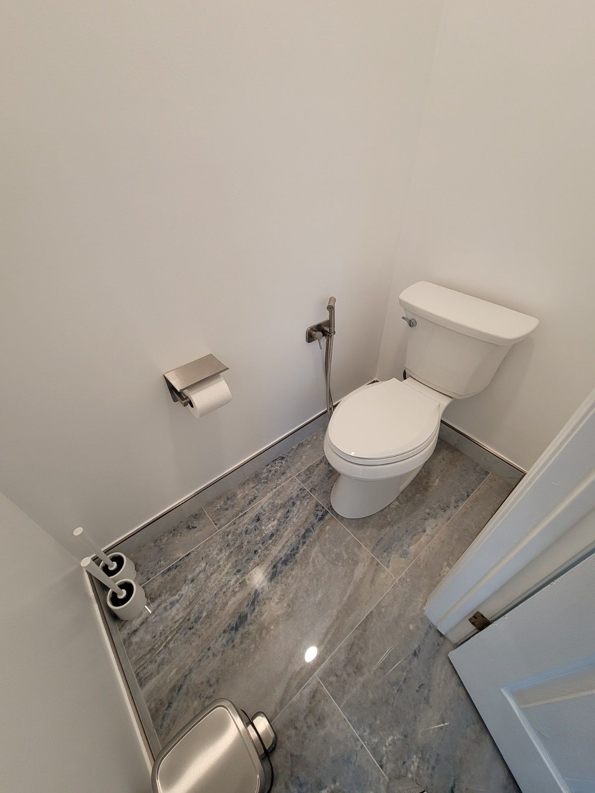 A modern white toilet with a handheld bidet sprayer, a wall-mounted toilet paper holder, and gray marble-look floor tiles.