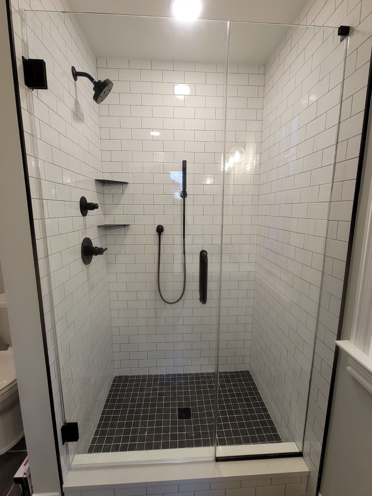 A modern shower with white subway tile walls, a dark gray patterned floor, and black fixtures enclosed by a glass door.