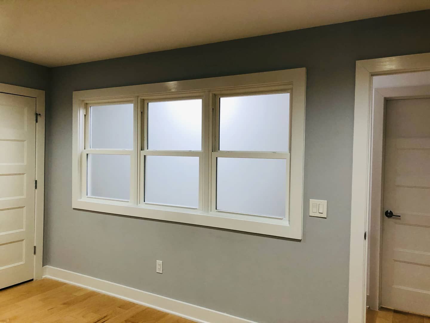 A room with gray walls, light wood floors, two white doors, and a three-section horizontal window with frosted glass.