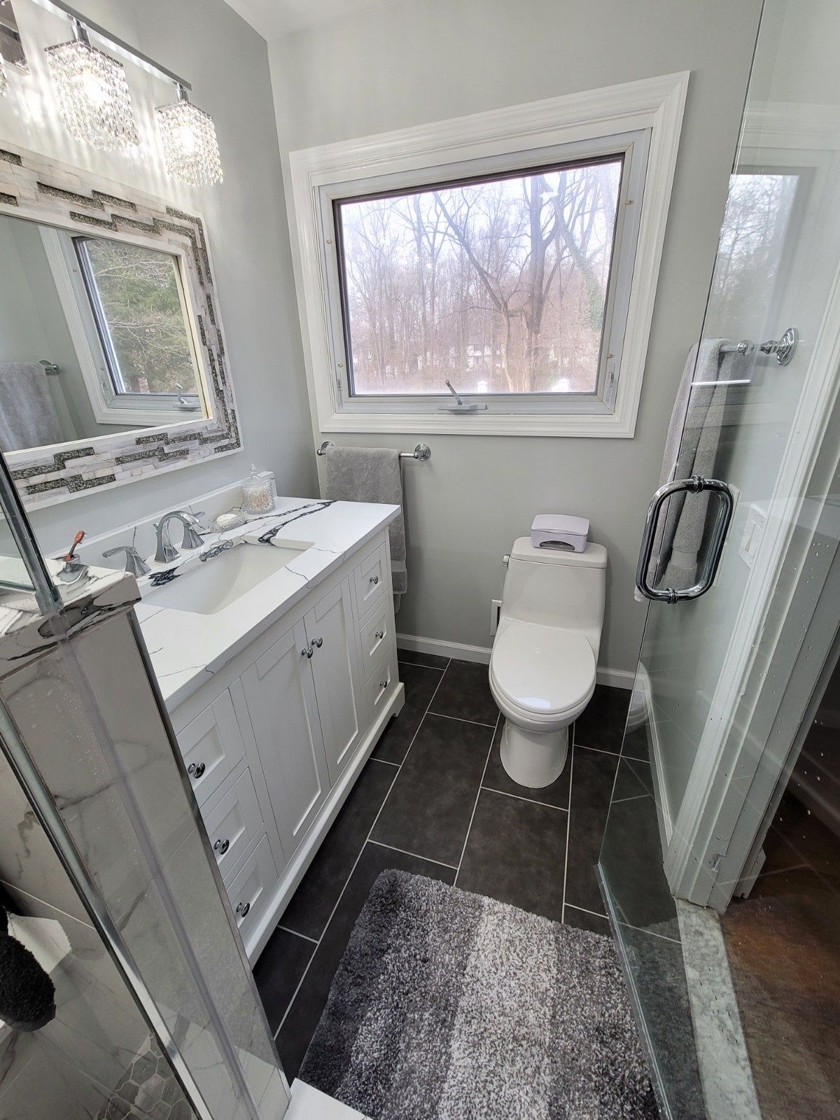 Modern bathroom with a white vanity, a light fixture, a window with a view of trees, and a toilet on a dark tile floor.