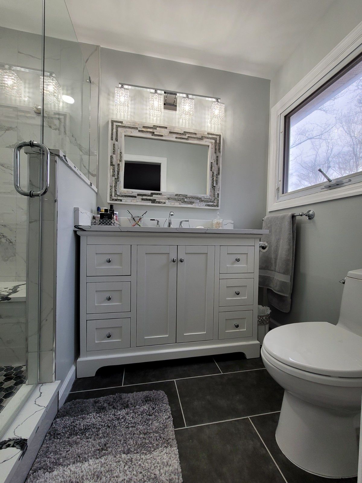 A modern bathroom features a white vanity with a framed mirror, a glass shower, dark tile floors, and a white toilet.