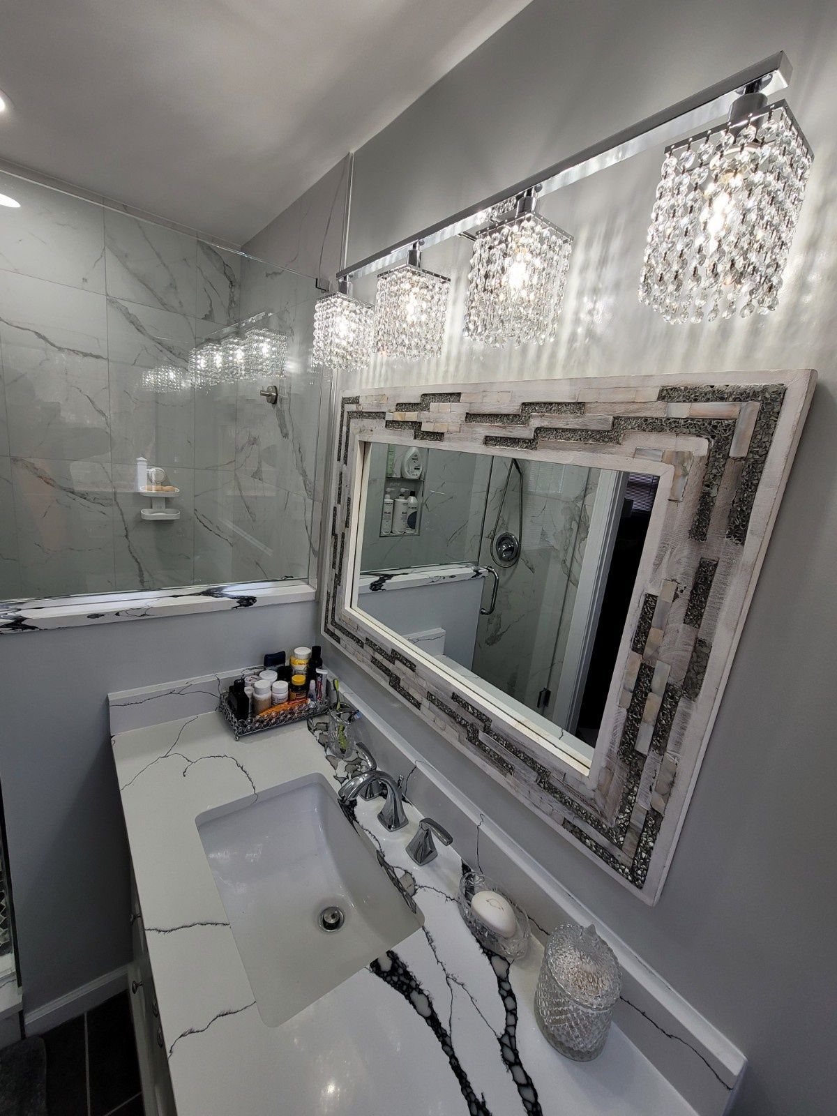 Modern bathroom vanity with marble countertop, decorative crystal light fixtures, and a textured white-framed mirror.