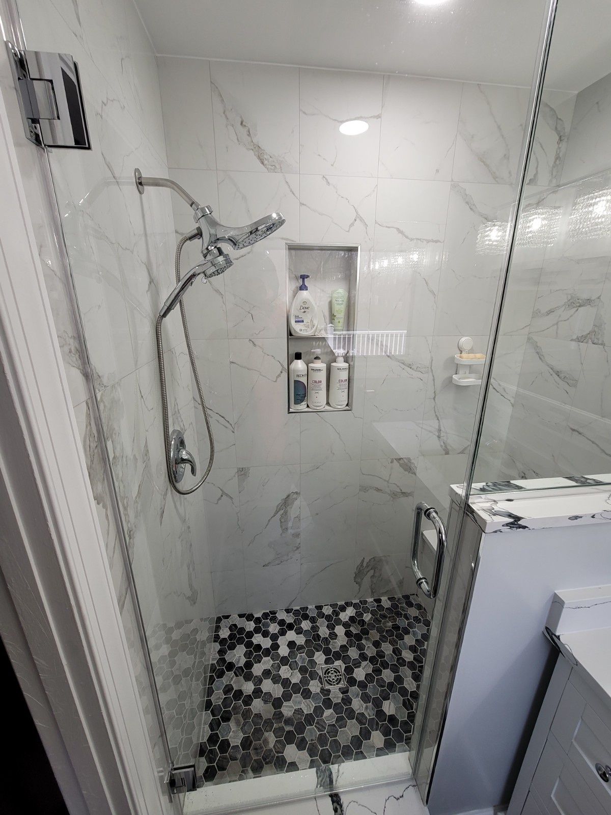 A tiled shower with a glass door, marble-patterned walls, a recessed niche with toiletries, and a hexagonal tile floor.