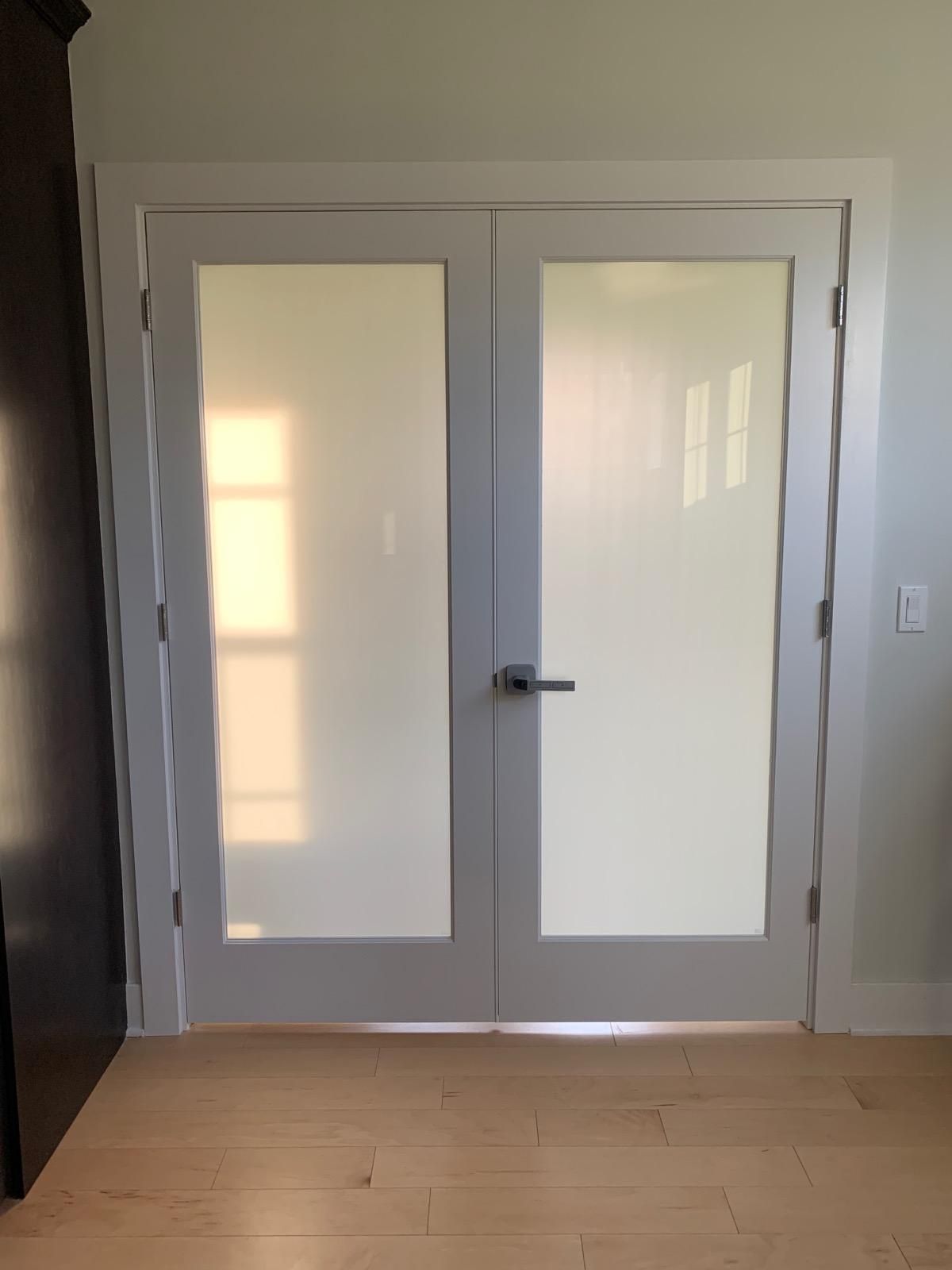 Double white French doors with frosted glass panes, featuring a modern silver handle, set in a light-colored interior room.