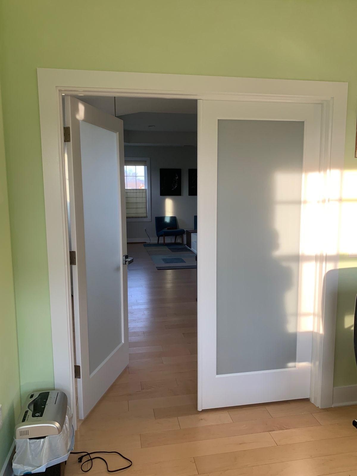 A doorway with white trim and frosted glass panels leads from a light green room into an open living space with wood floors.
