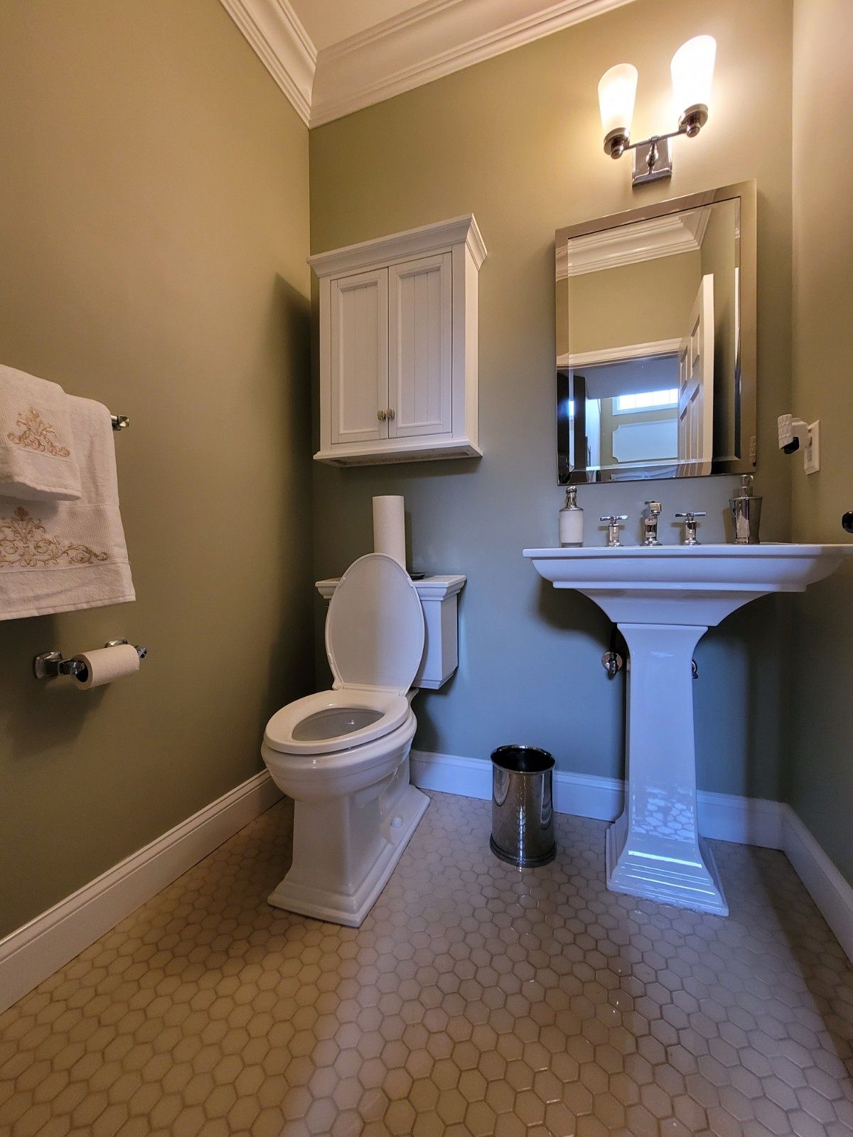 A bathroom with olive walls, a white pedestal sink, a white toilet with a wall cabinet above it, and hexagon tile floors.