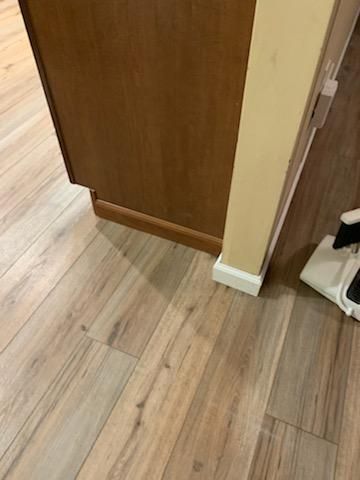 A wooden cabinet base meets a white baseboard and a wall corner on light wood-grain flooring.