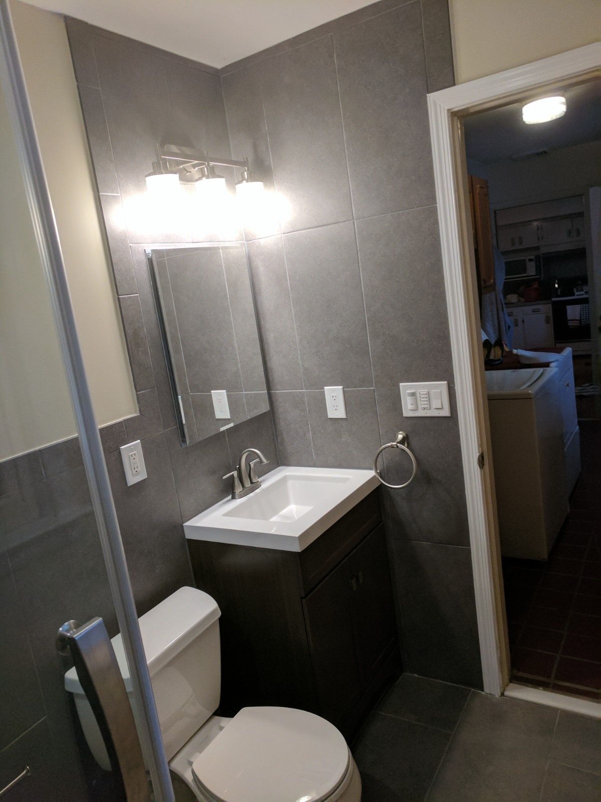 A bathroom with grey tiled walls, a small vanity with a white sink, a toilet, a mirror, and a wall-mounted light fixture.