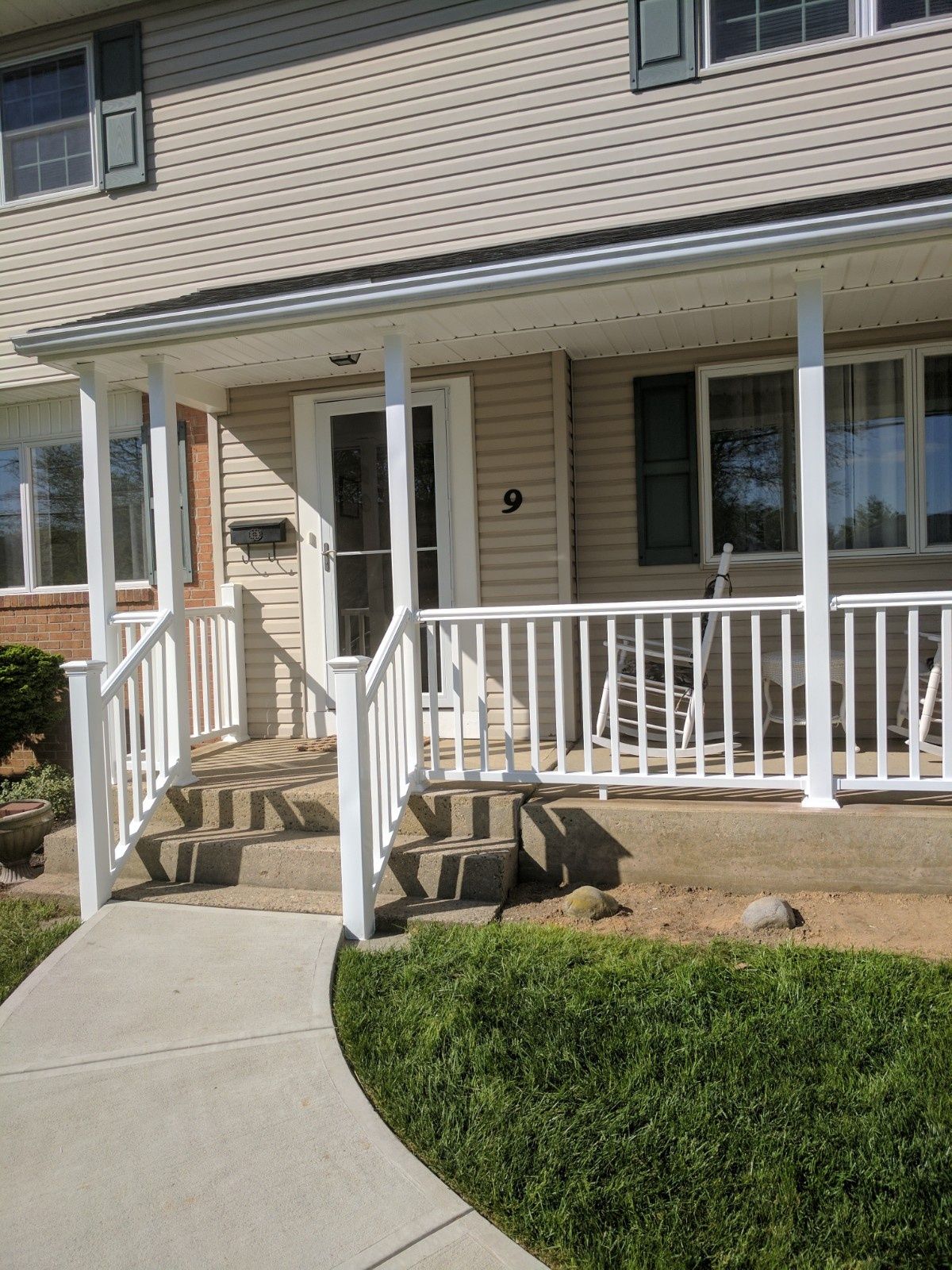 A front entrance to a beige house with a white porch railing, concrete steps, and a sidewalk leading to the door.
