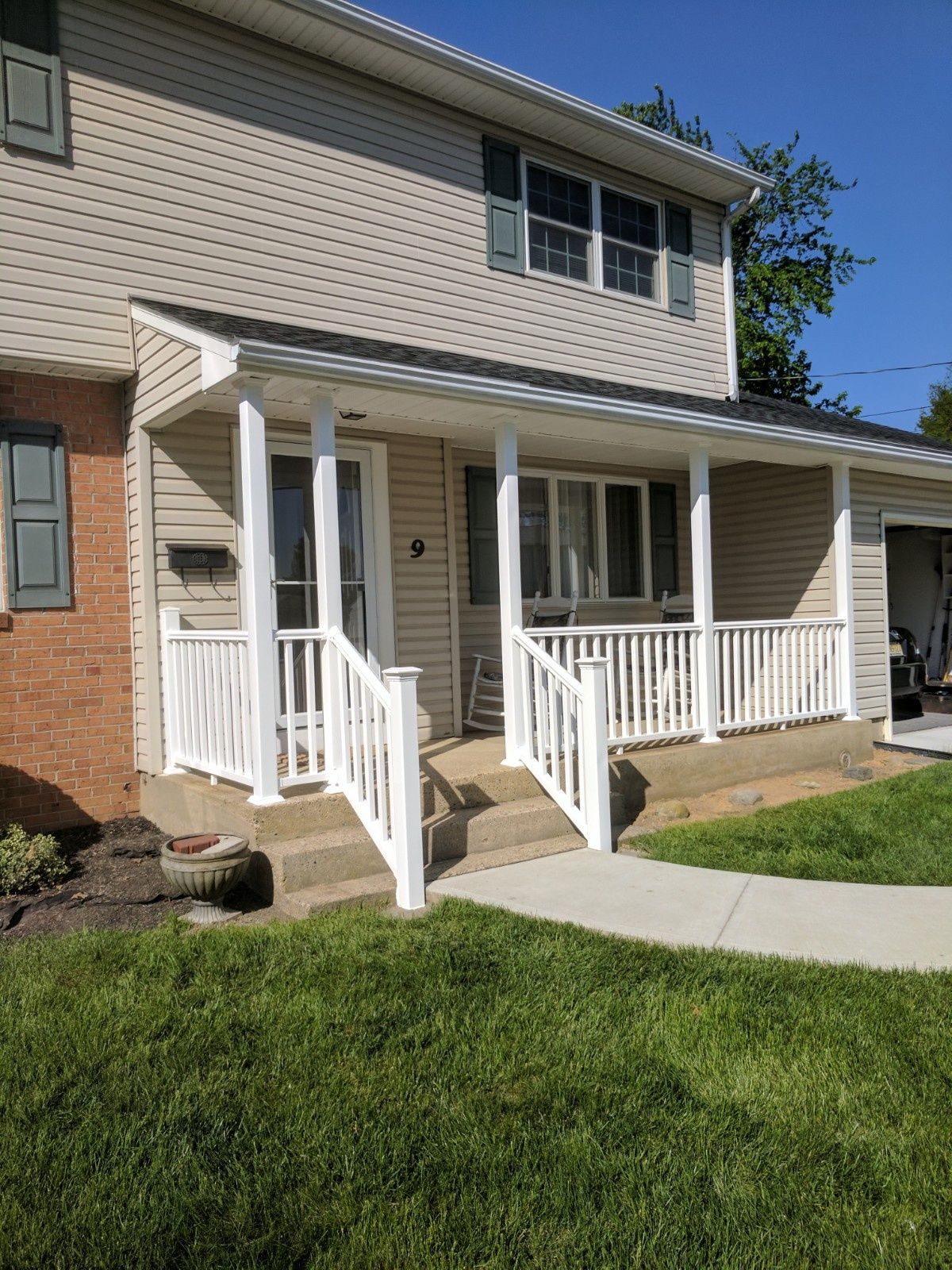 A two-story tan vinyl-sided house with a white front porch, stairs, and railing under a clear blue sky.