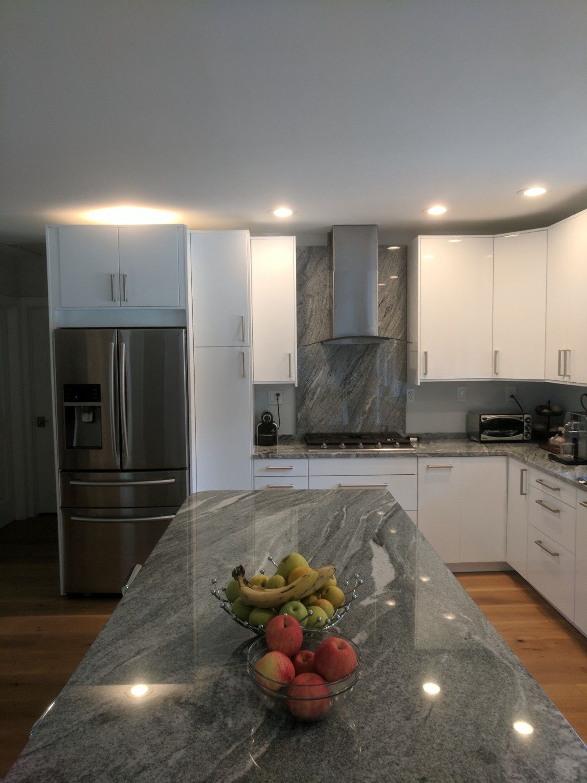 A modern kitchen with white cabinets, stainless steel appliances, and a gray granite-topped island holding a bowl of fruit.
