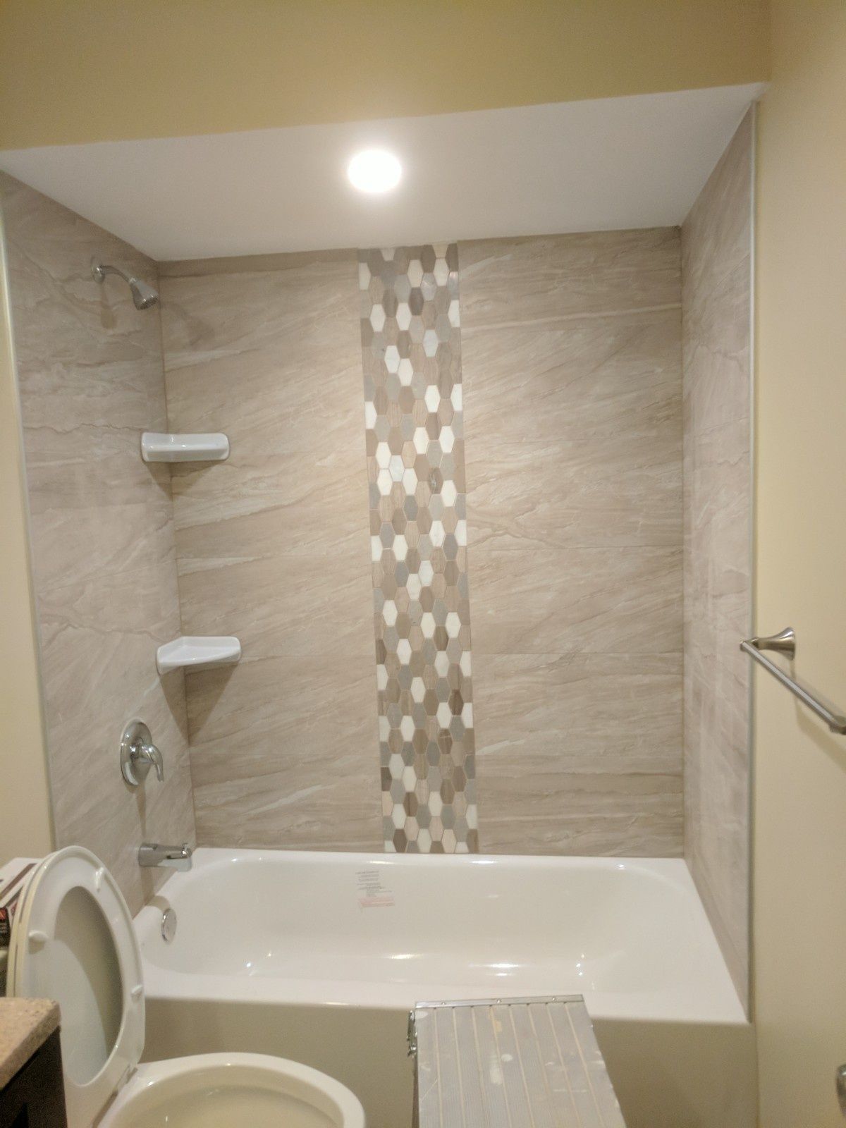A bathtub and shower alcove with light wood-look tile walls, a vertical mosaic accent stripe, and two corner shelves.