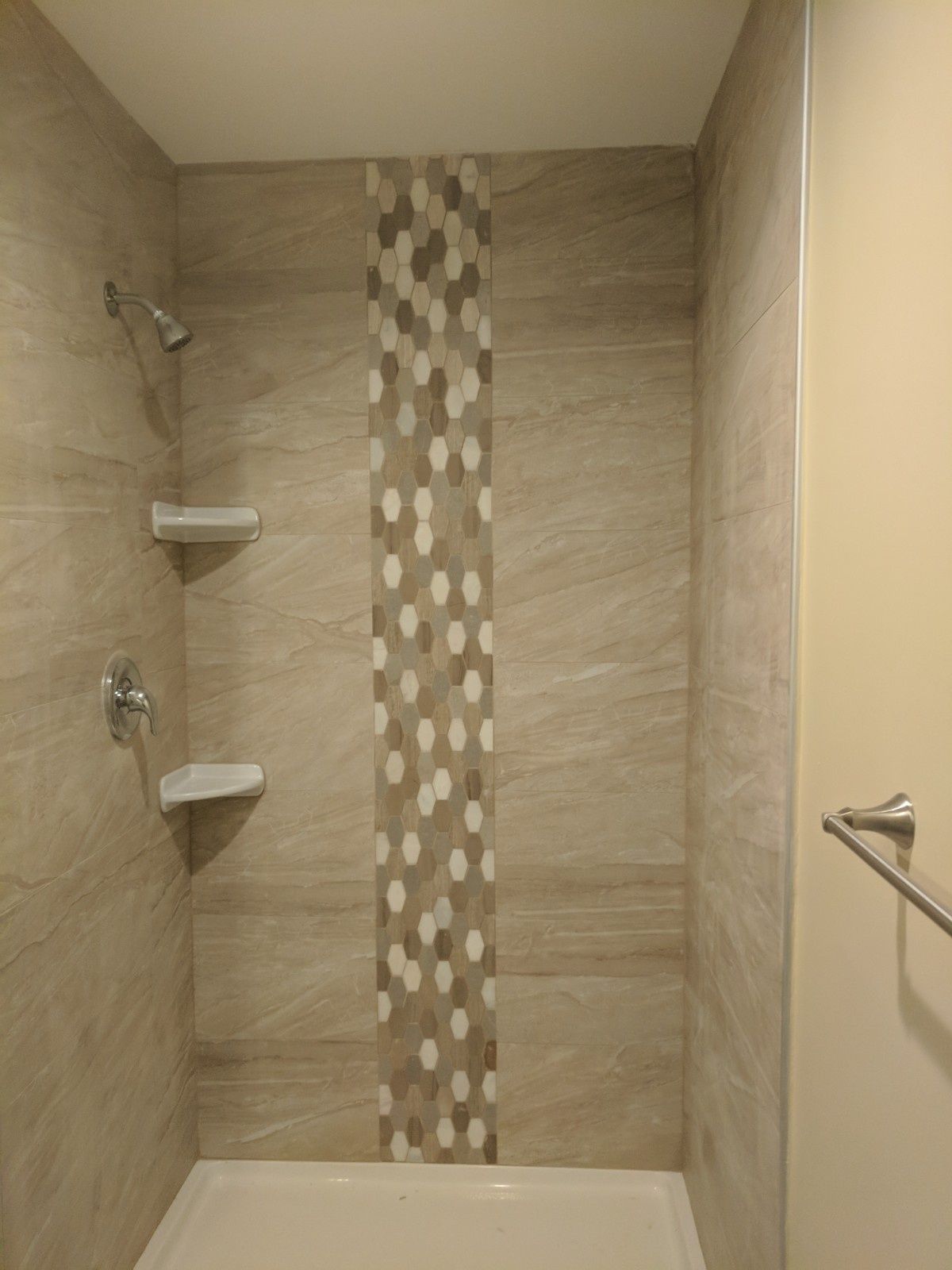 A shower with light brown, marble-look tiles, a central vertical column of mosaic tiles, and two built-in corner shelves.