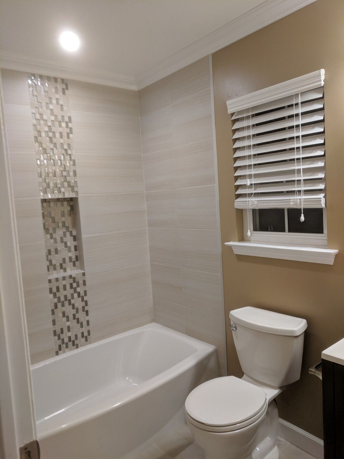 A modern bathroom features a white bathtub with a tiled niche and a toilet beneath a window with white horizontal blinds.