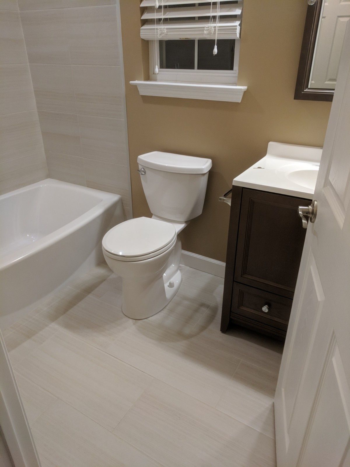 A small bathroom with light tile floors, a white tub, a white toilet, and a dark wood vanity with a white countertop.