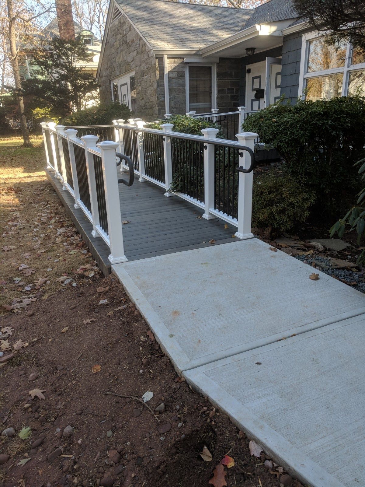 A ramp with gray decking and white railings leads to the entrance of a stone house with a concrete walkway.