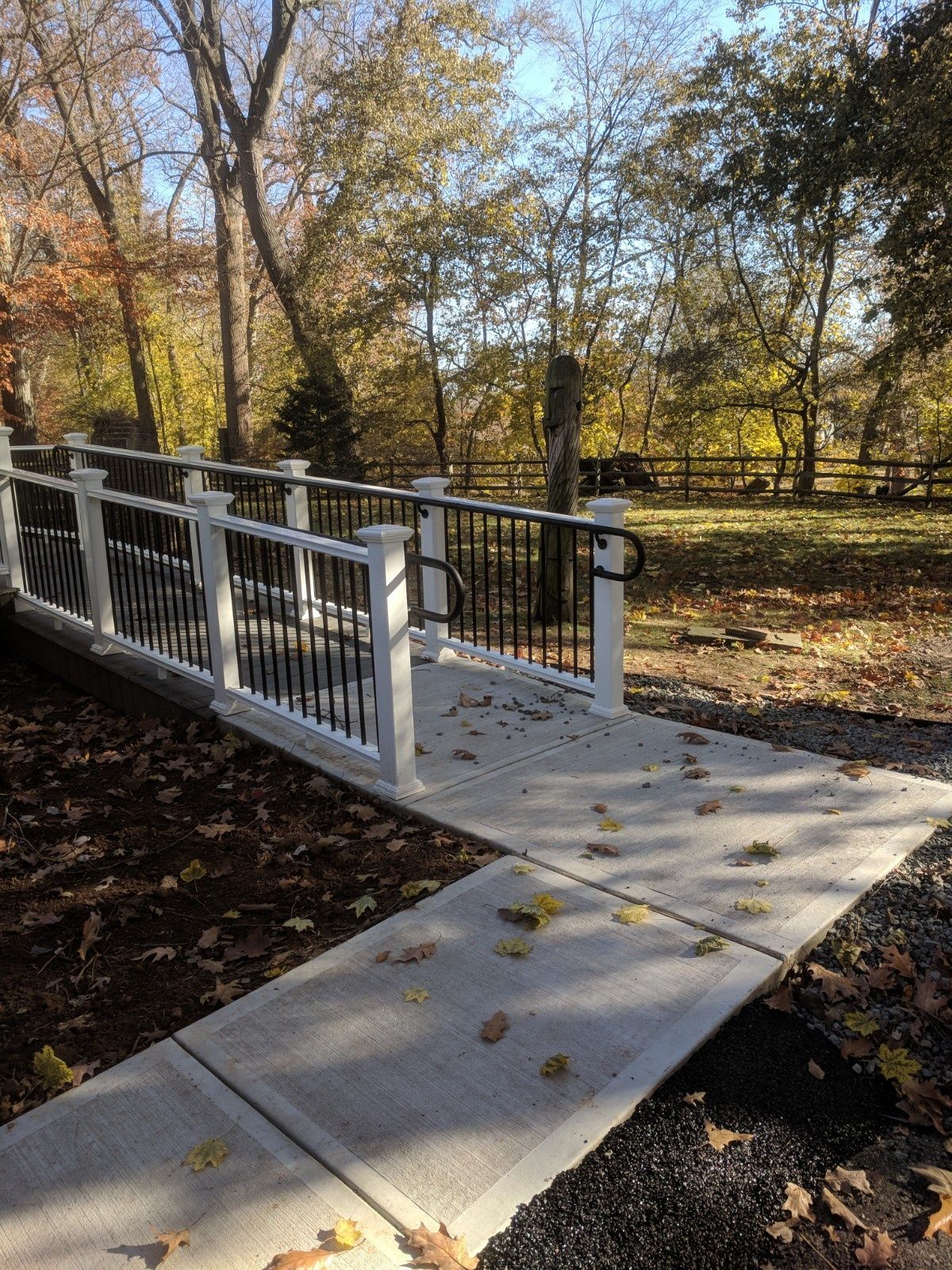 A concrete wheelchair ramp with white posts and black railings, leading downward toward a wooded area in autumn.