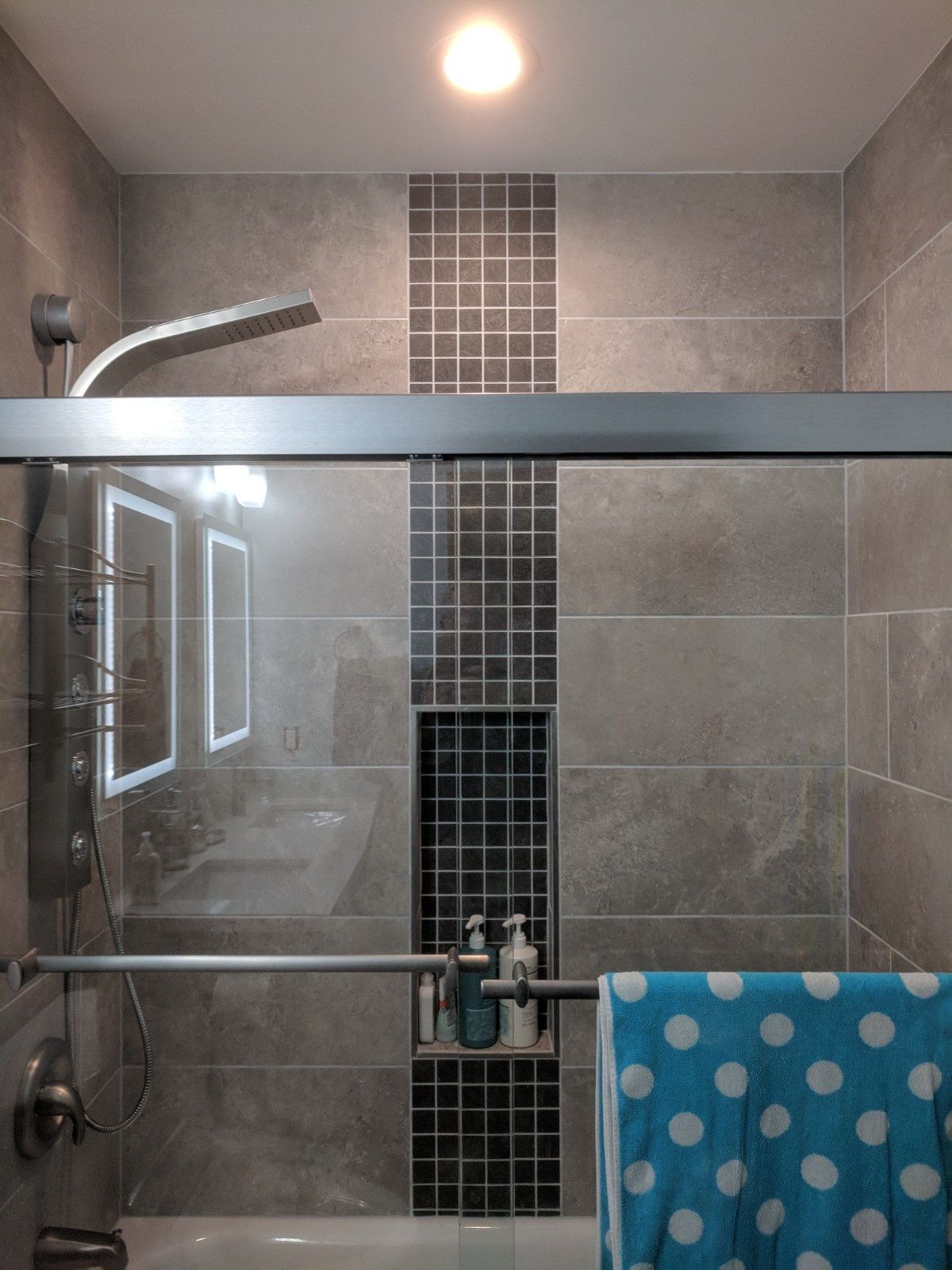 Modern shower with gray stone tiles, a dark mosaic accent wall, a glass door, and a blue polka-dot towel on the handle.