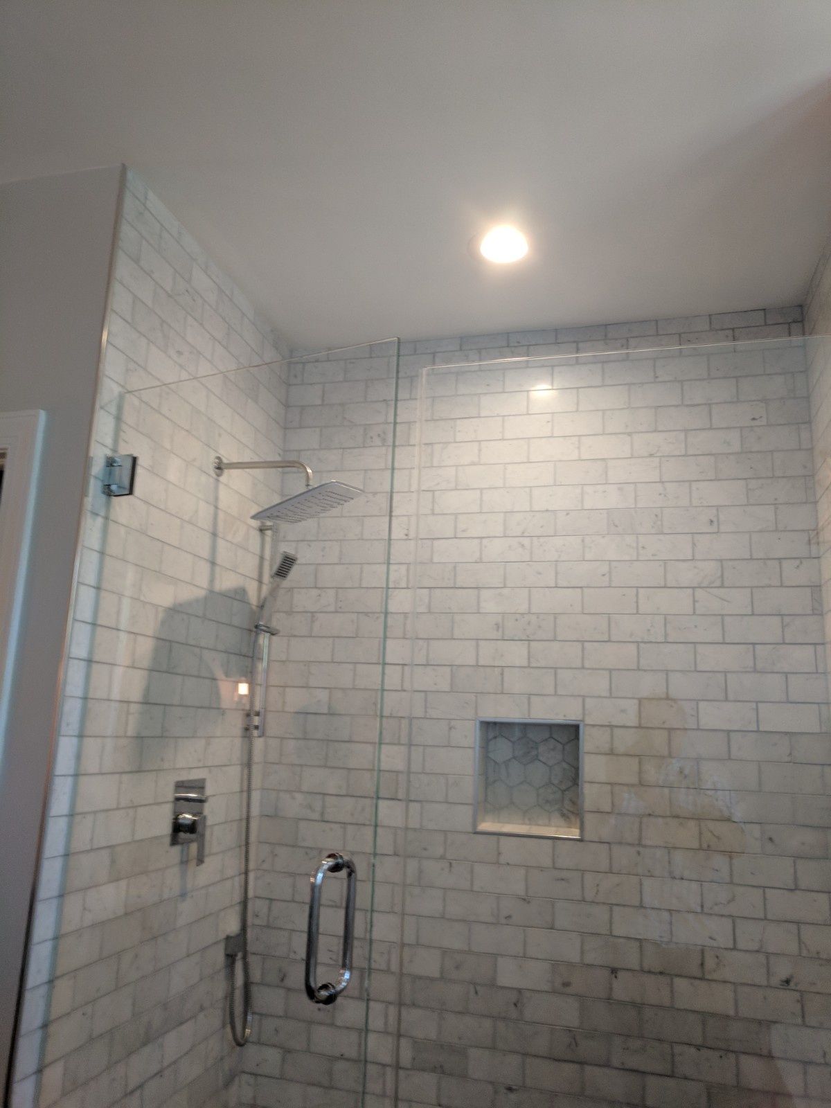A walk-in shower with light-colored brick-style wall tiles, a glass door, a silver rainfall showerhead, and a small niche.