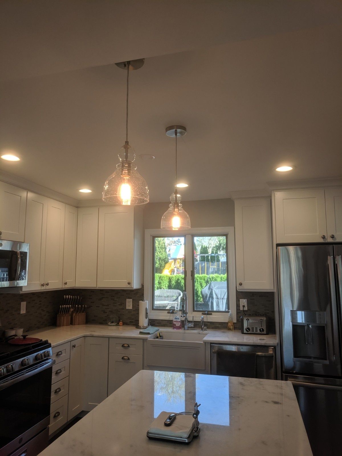A bright kitchen featuring white cabinets, stainless steel appliances, a marble-topped island, and two pendant lights.