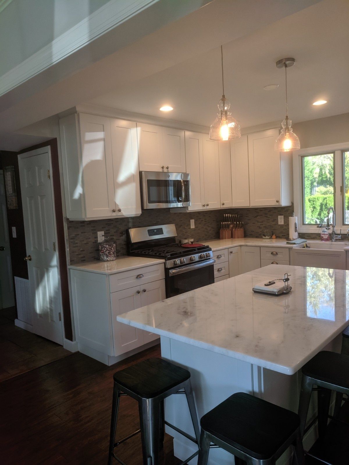 A white kitchen with a marble-topped island, dark bar stools, stainless steel appliances, and a window above the sink.