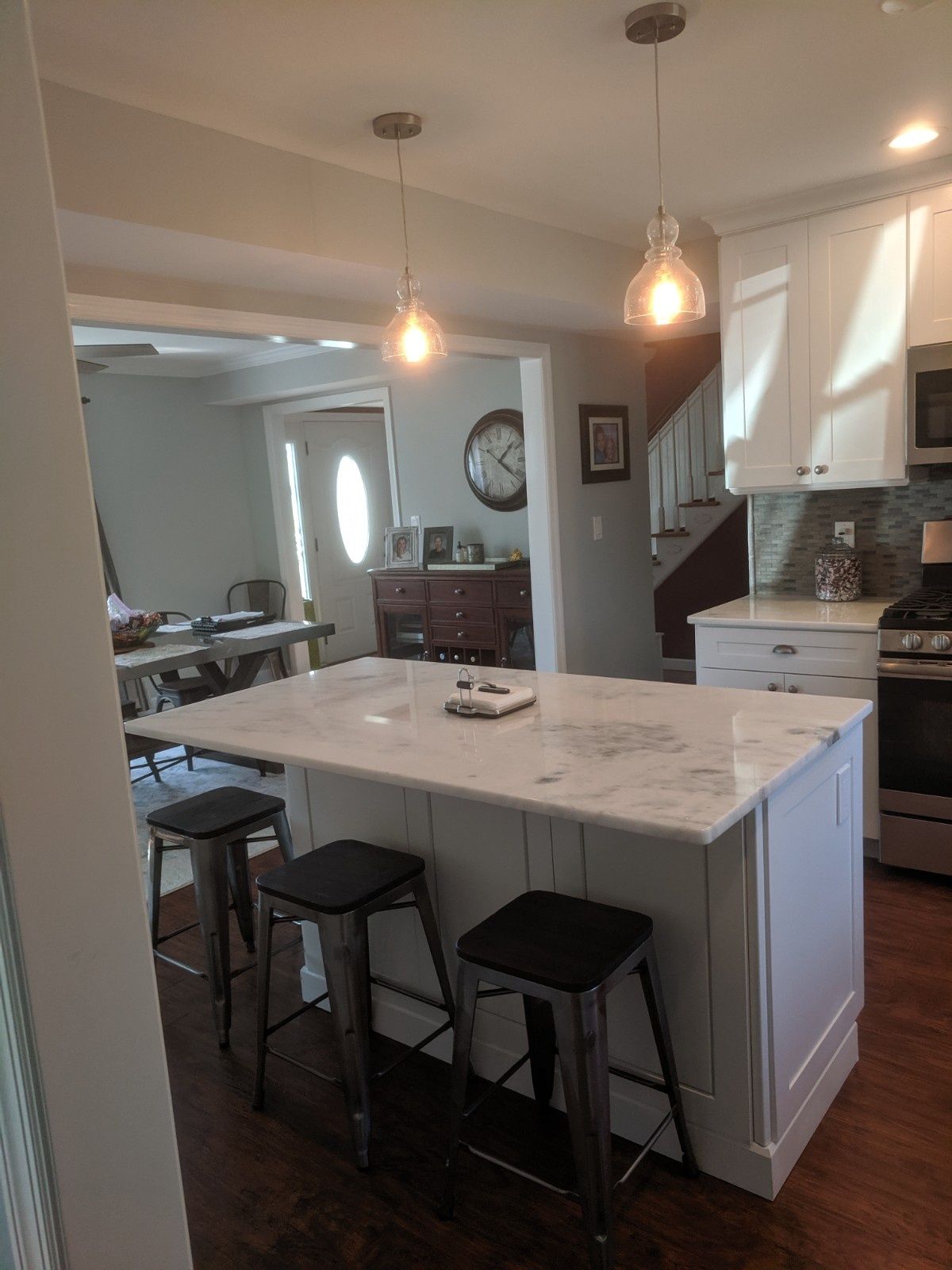 A kitchen island with three metal stools, marble countertop, and pendant lights, opening to a dining area.
