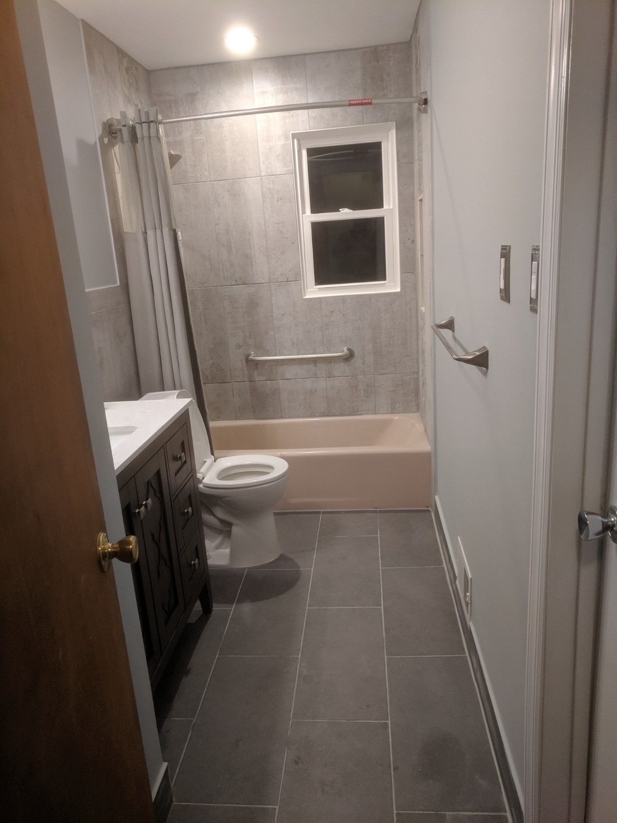 A narrow bathroom with gray tile floors, a dark vanity, a toilet, and a bathtub with gray wall tiles and a window.