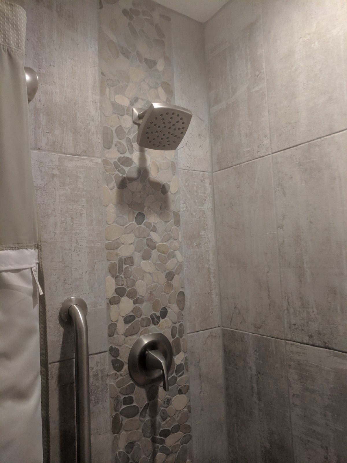 A shower stall with gray tile walls, a vertical accent strip of river stones, a silver shower head, and a metal grab bar.