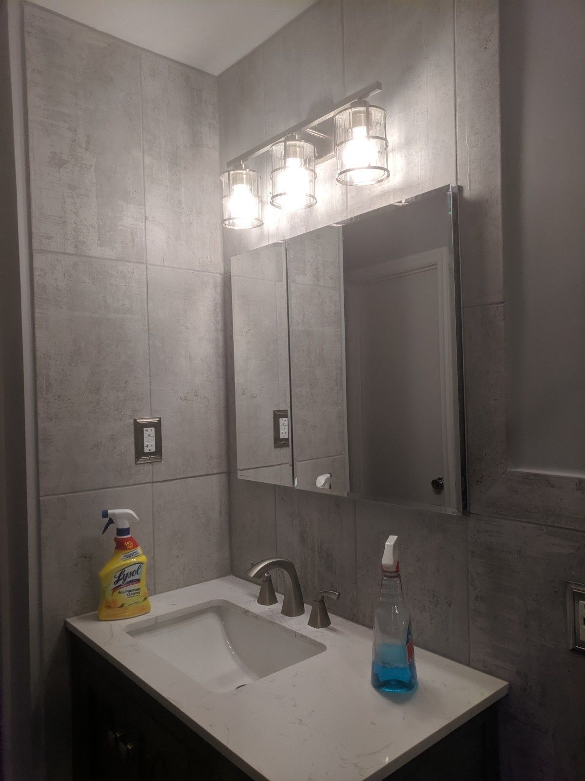 A modern bathroom vanity with gray tile walls, a white countertop, sink, medicine cabinet, and a three-light fixture.