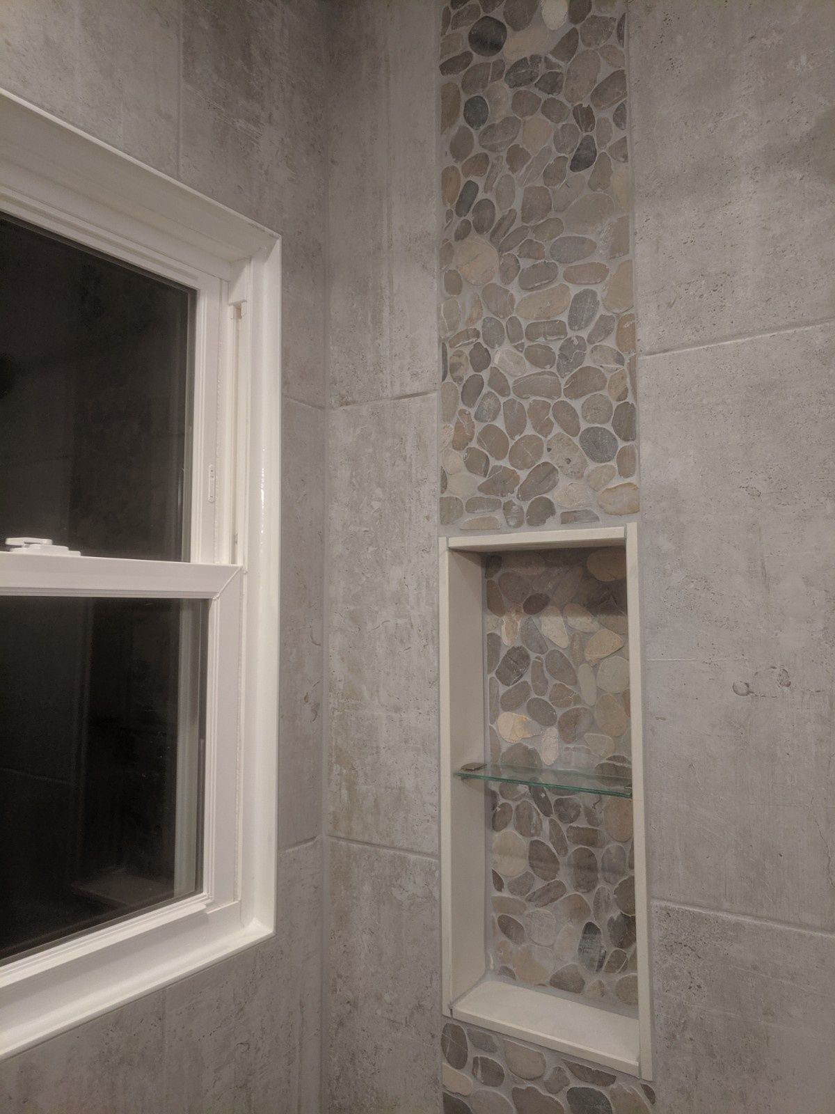 A tiled shower wall featuring a white-framed window next to a vertical strip of stone pebbles and a built-in recessed shelf.