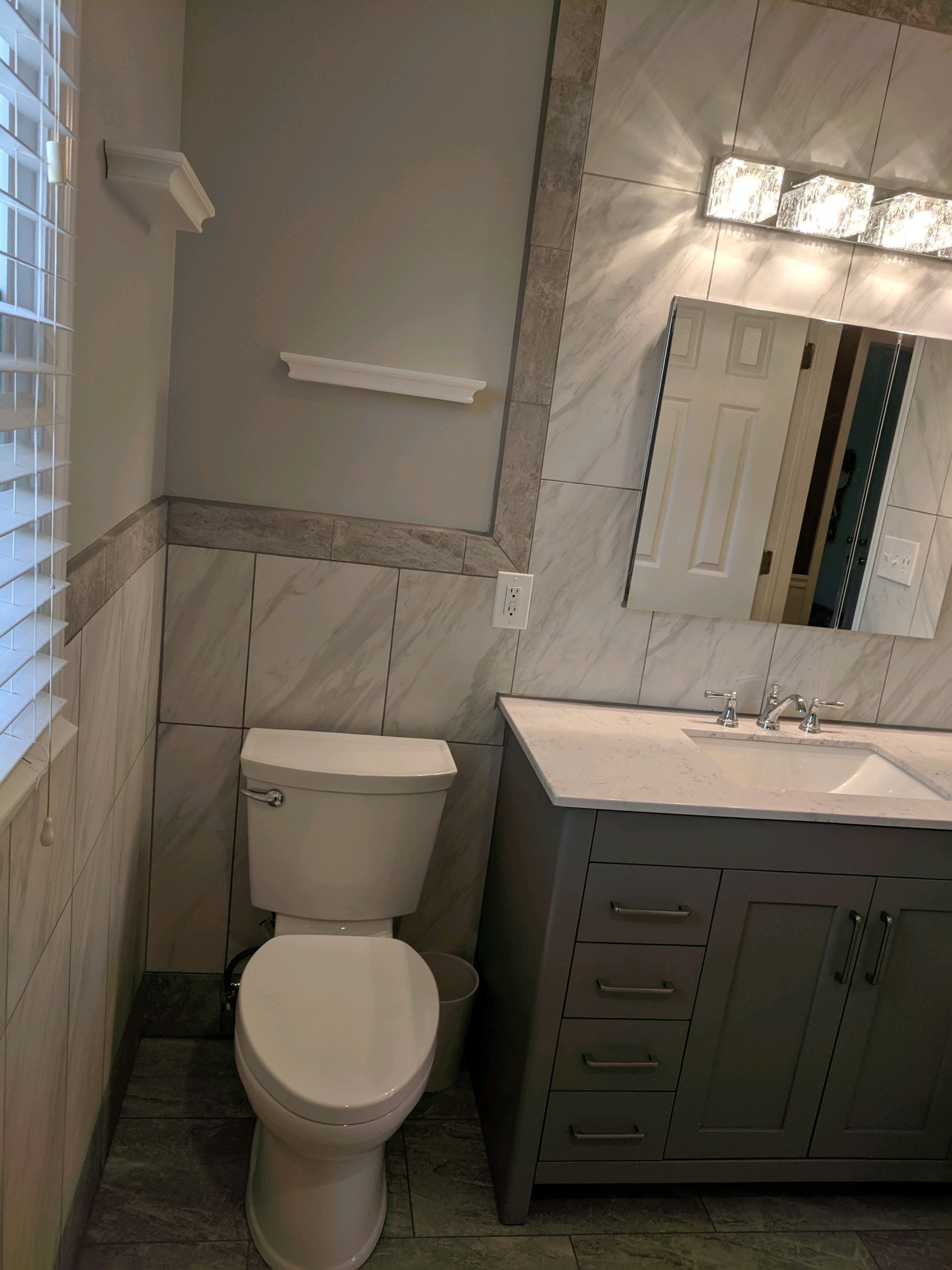 A gray bathroom vanity with a white sink, a mirror with lights above, and a white toilet next to a tiled wall.