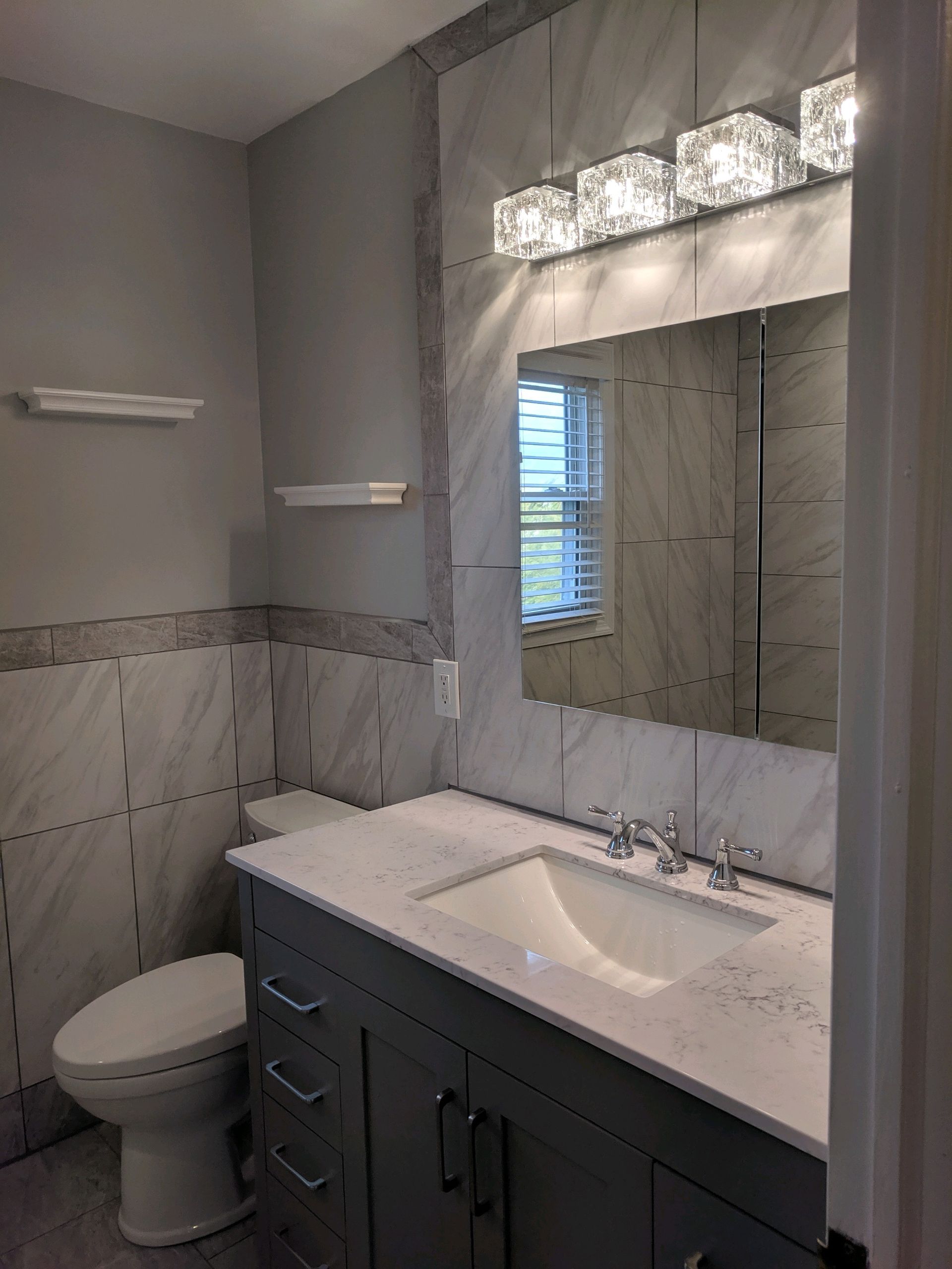 A modern bathroom with grey walls, white marble-style tiled walls, a dark grey vanity, white countertop, and glass lighting.