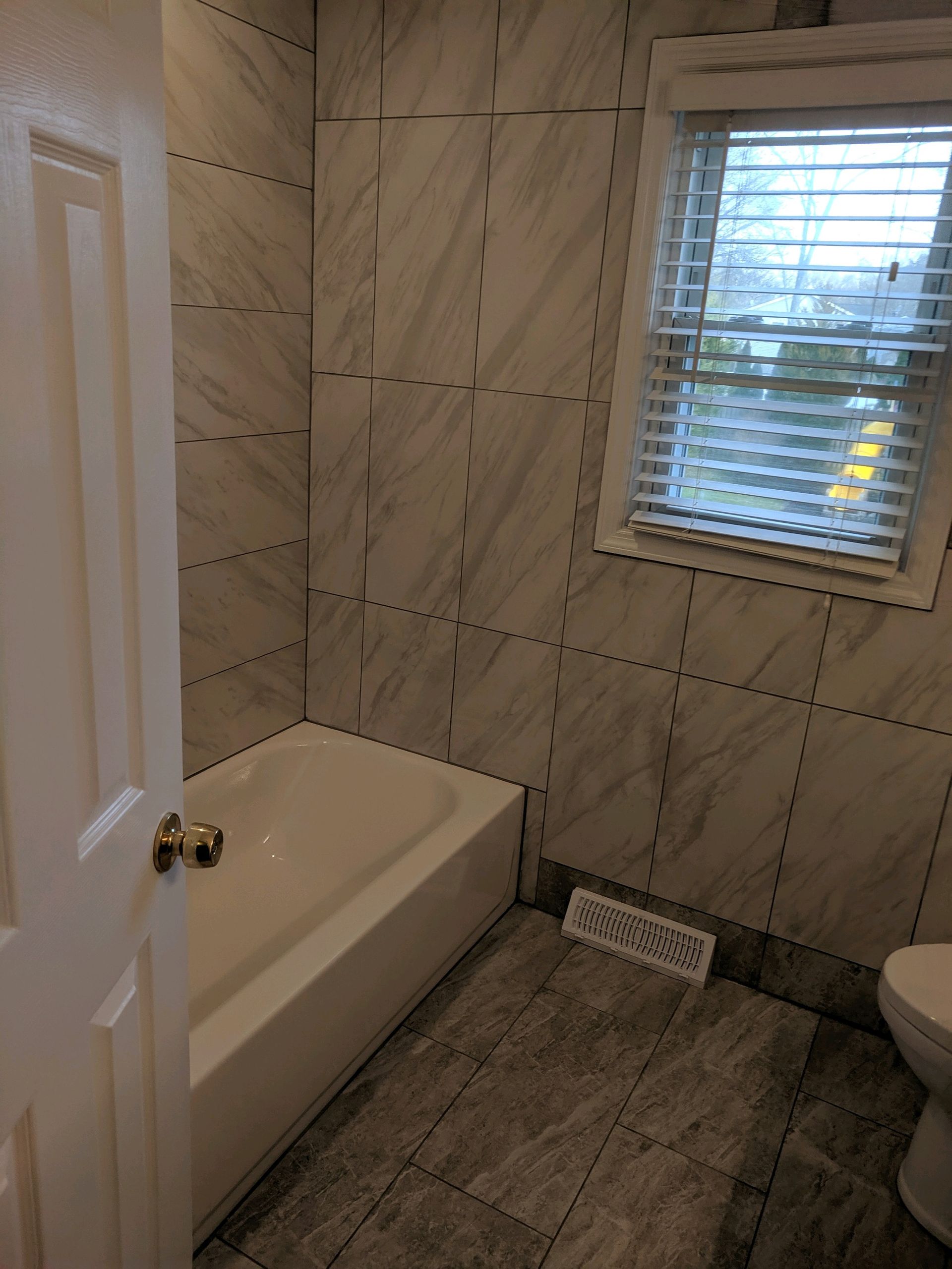 A bathroom with a white bathtub, gray marble-patterned tiled walls, gray tiled floor, and a window with white blinds.