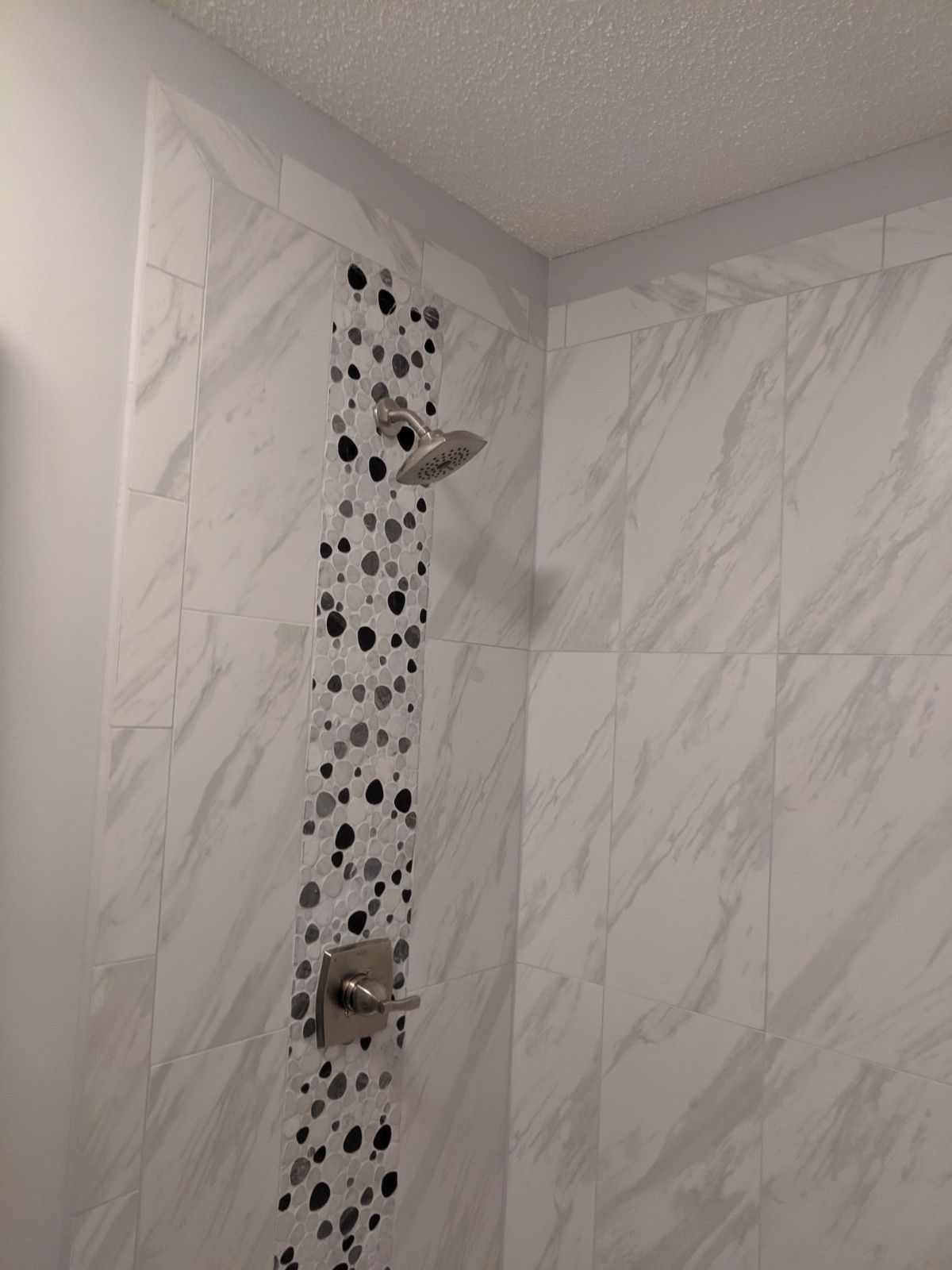 A shower wall with large white marble-look tiles and a vertical accent strip of black and white speckled mosaic tiles.